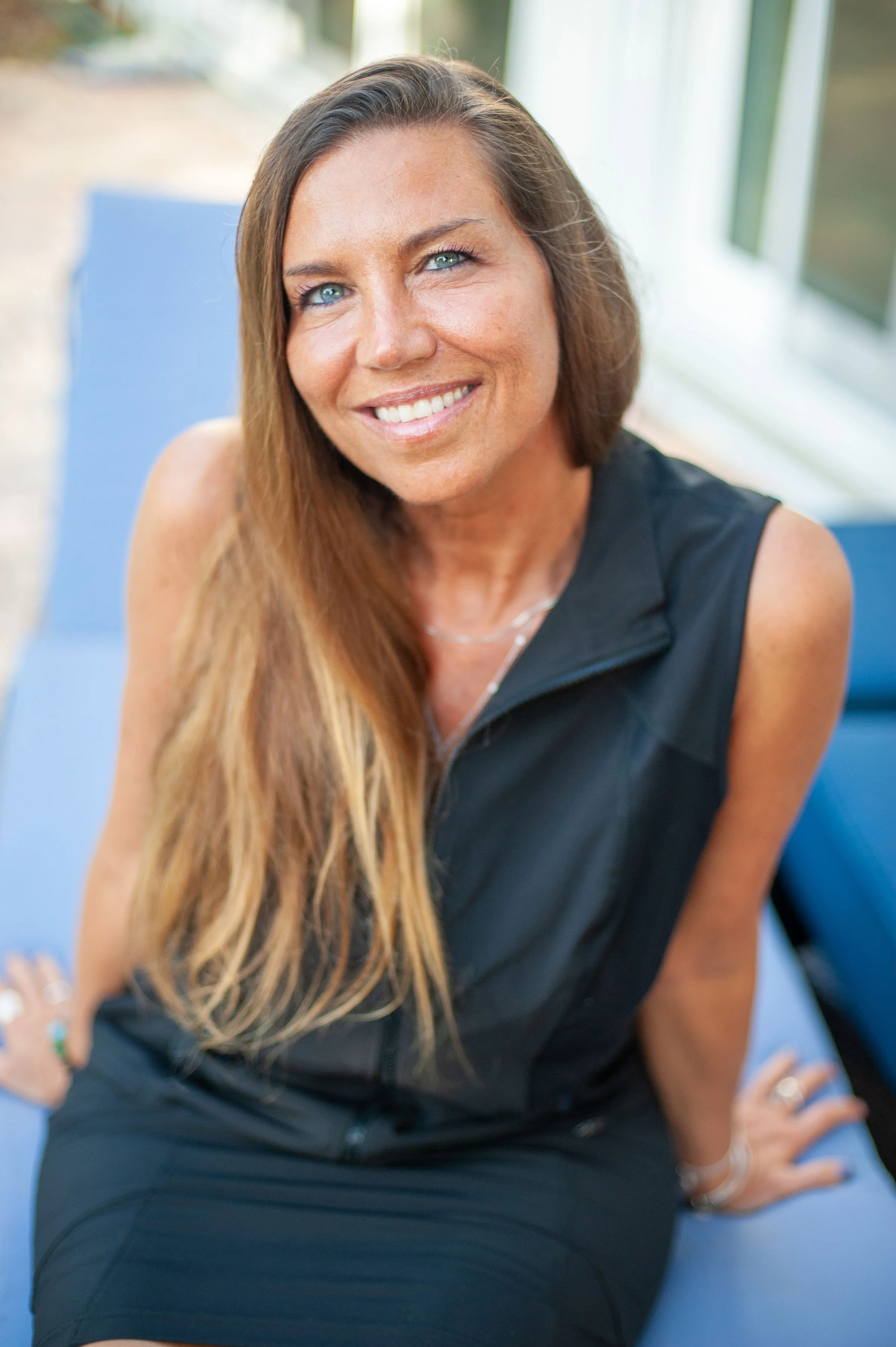 A woman with long brown hair and bright blue eyes sitting outdoors on a blue lounge chair, smiling at the camera.