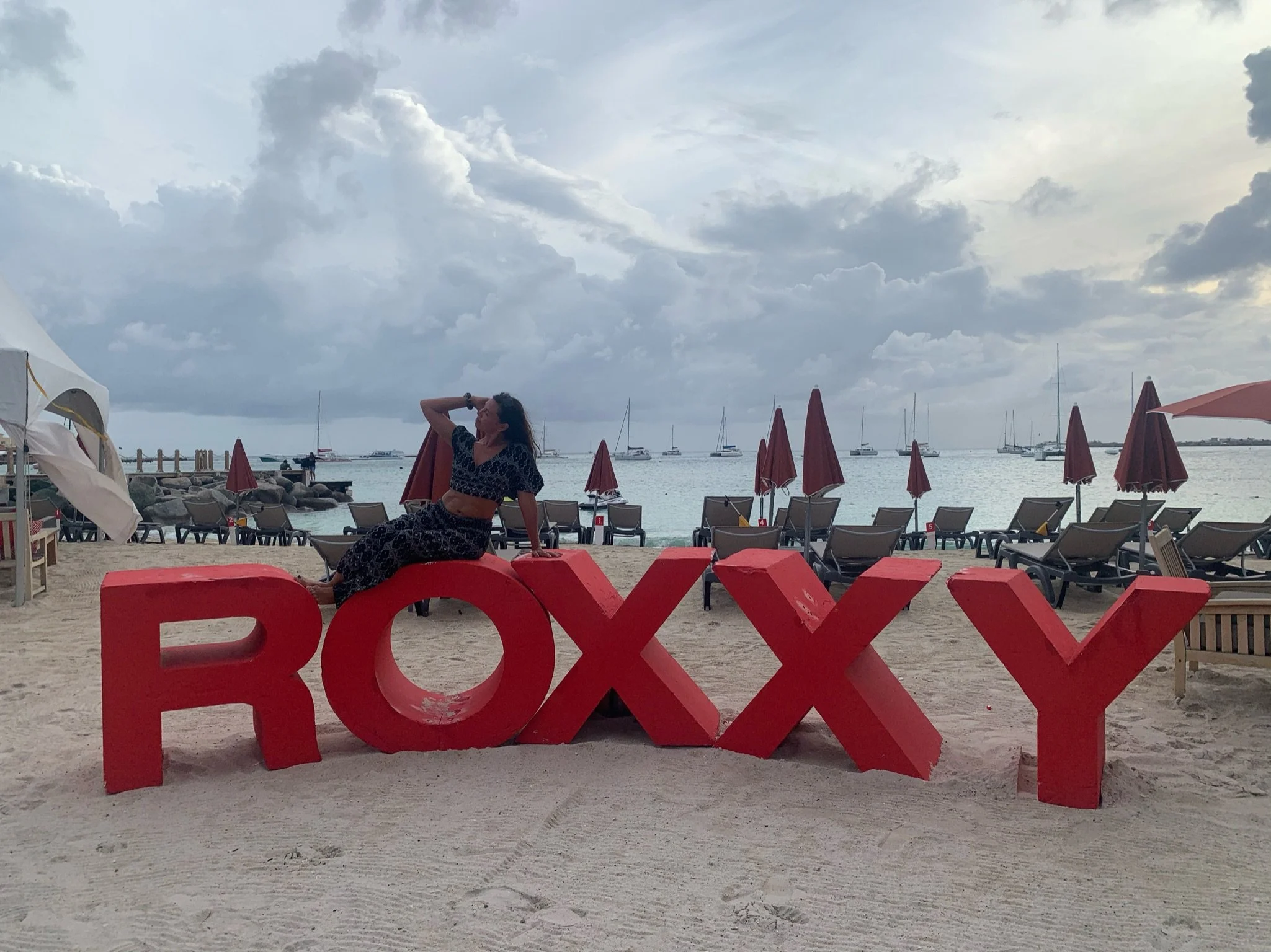 A woman in a black patterned outfit sitting on a large red 'ROXXY' sign on a sandy beach with beach chairs and umbrellas in the background, and boats on the water under cloudy sky.
