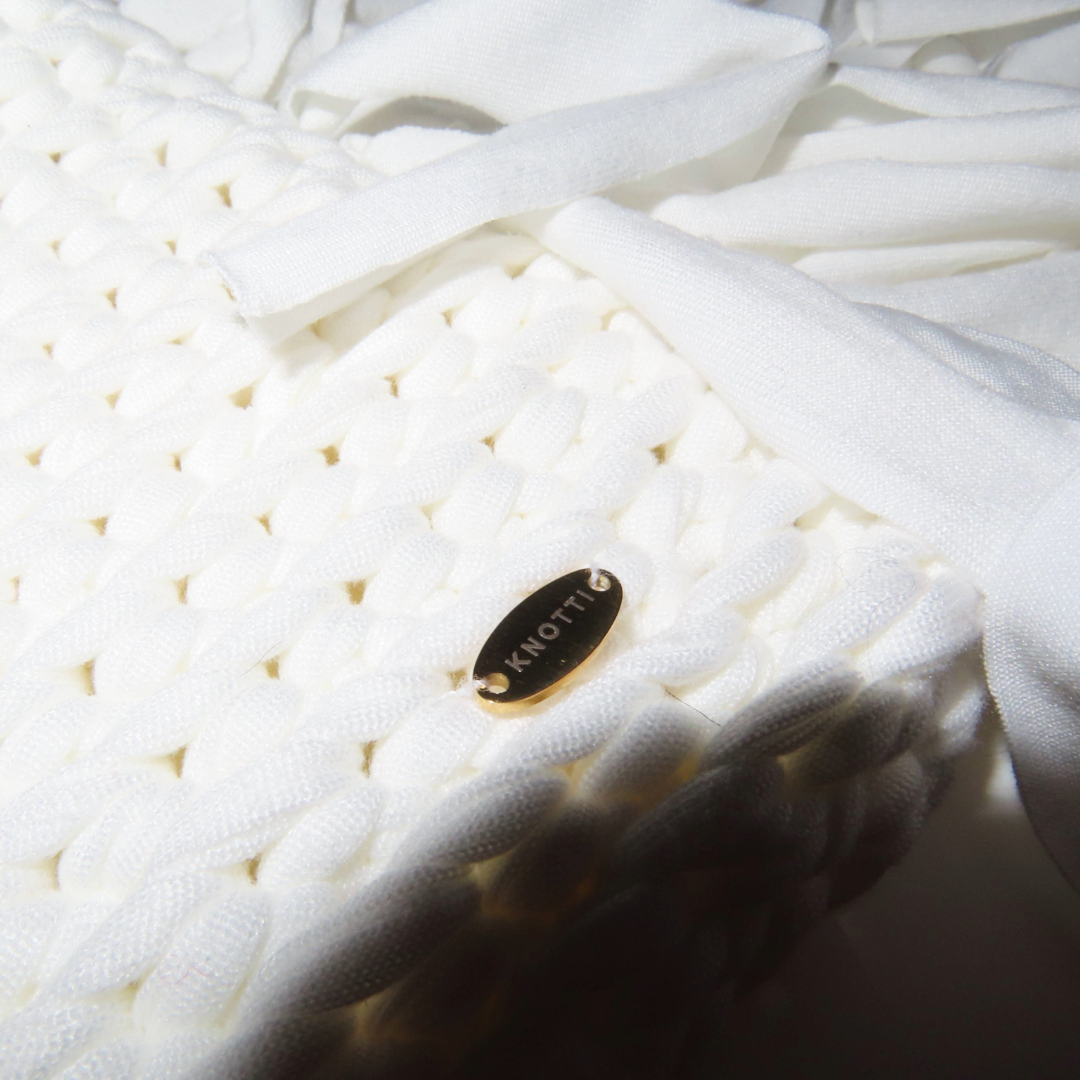 Close-up of a white knit fabric with a black oval tag labeled 'KNOTT' and white ribbons resting on the surface.
