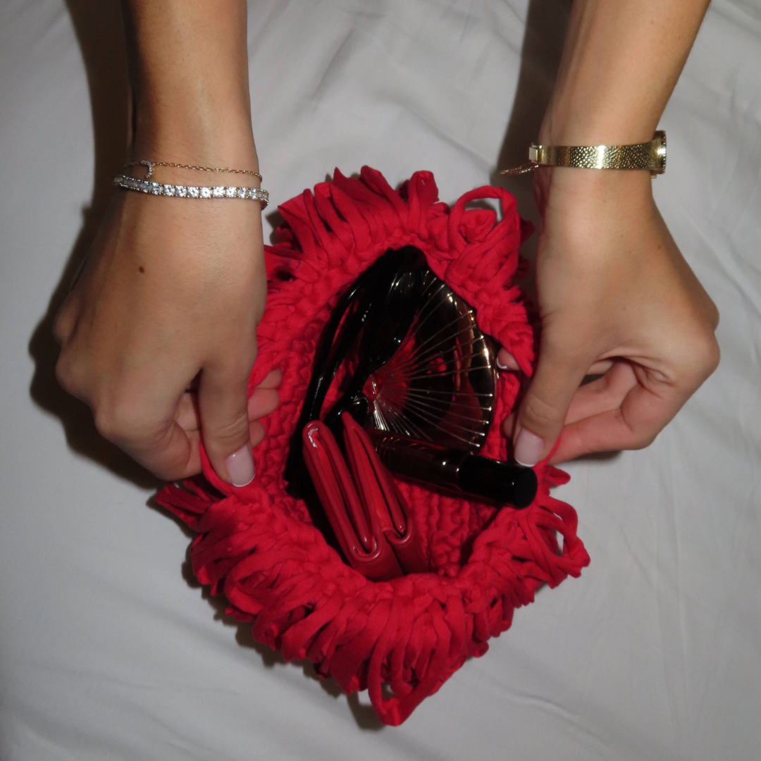 Hands holding a red cloth bag containing black and red makeup tools, on a white surface.