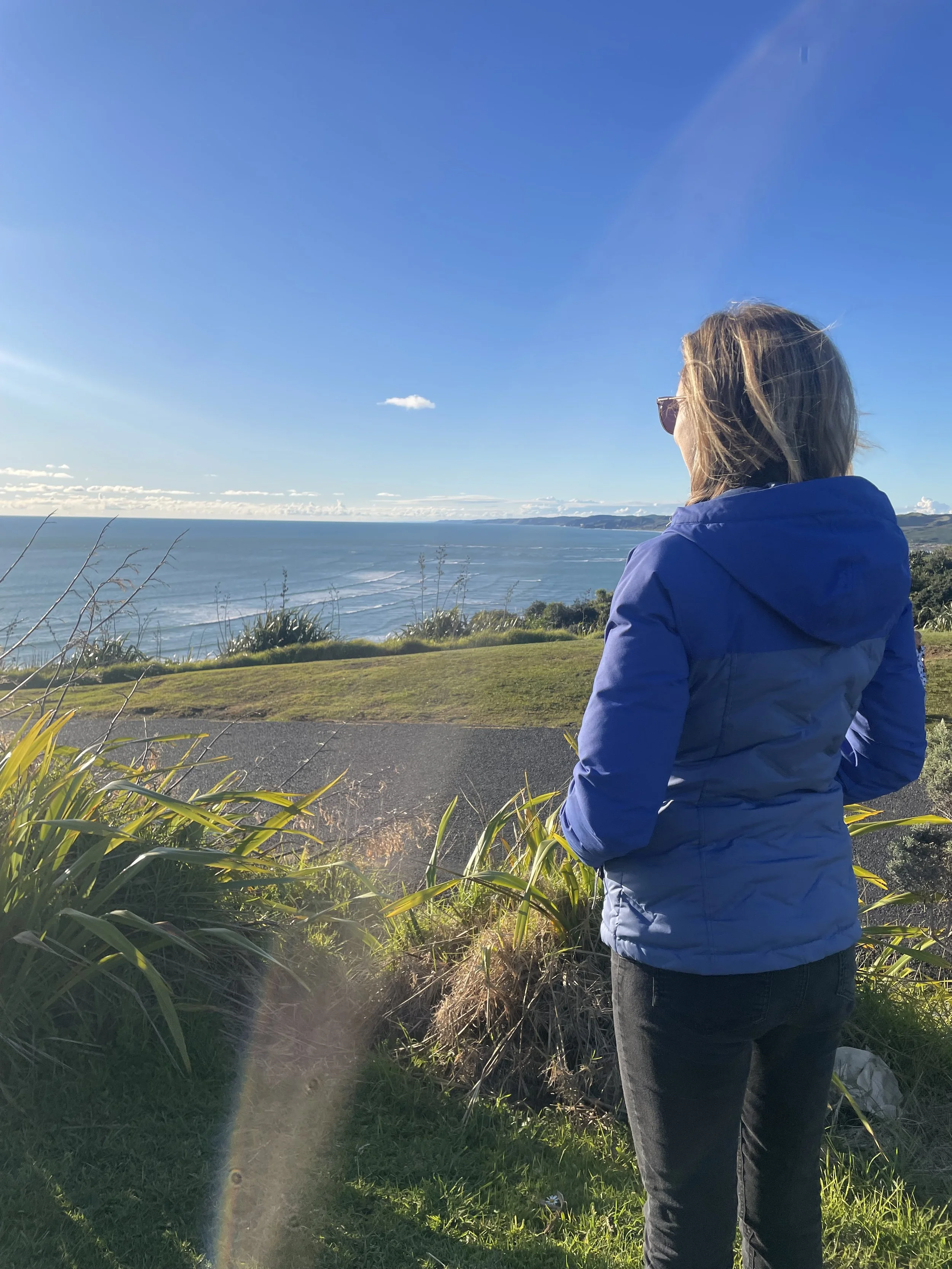 Woman in blue jacket and sunglasses looking at the ocean from a grassy cliffside on a sunny day.