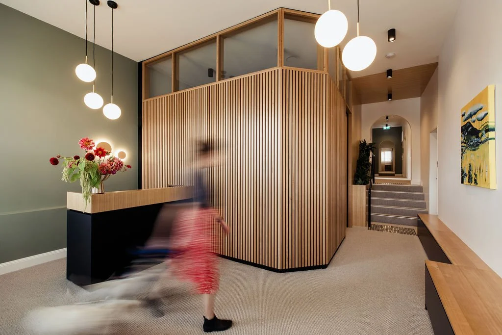 Modern interior lobby with wooden wall paneling, pendant lights, a bench, and artwork on the walls. A blurred child is walking through the space.