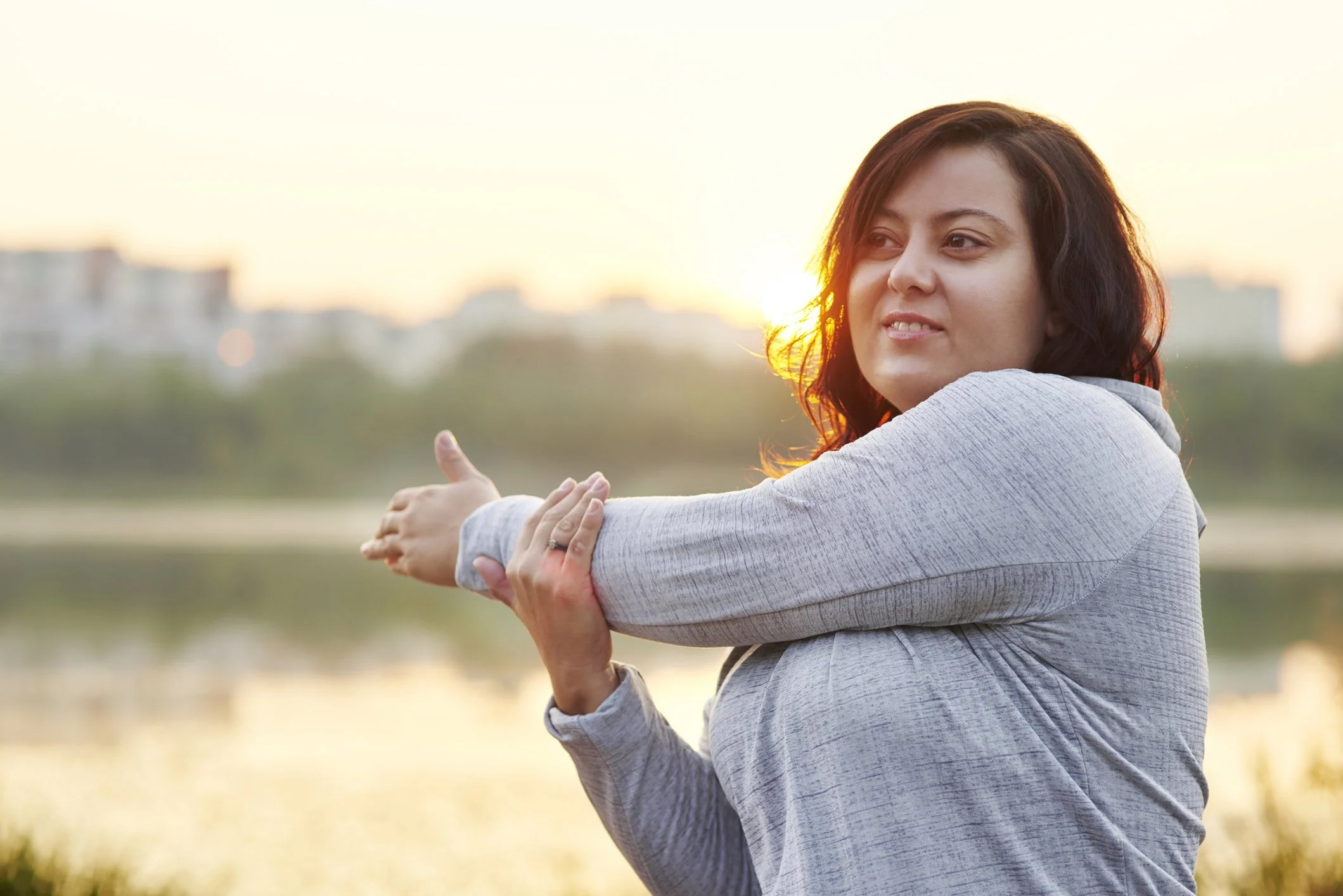 Woman stretching by a body of water during sunset.