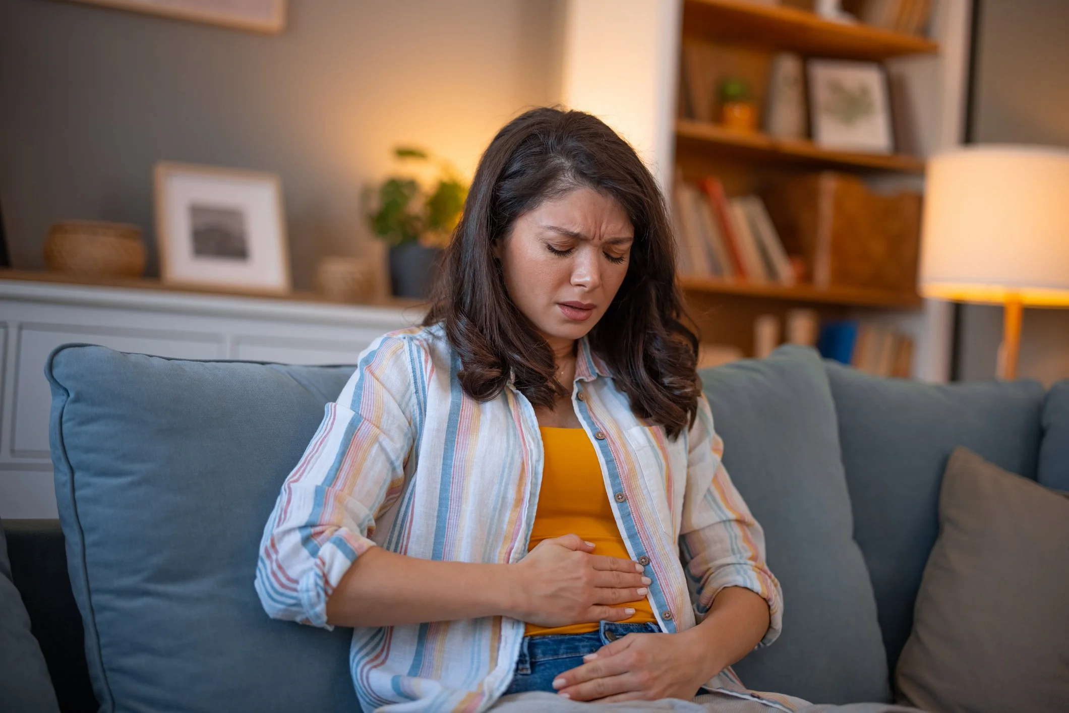 A woman sitting on a sofa clutching her stomach in pain, indicating discomfort or stomach ache, in a cozy living room.