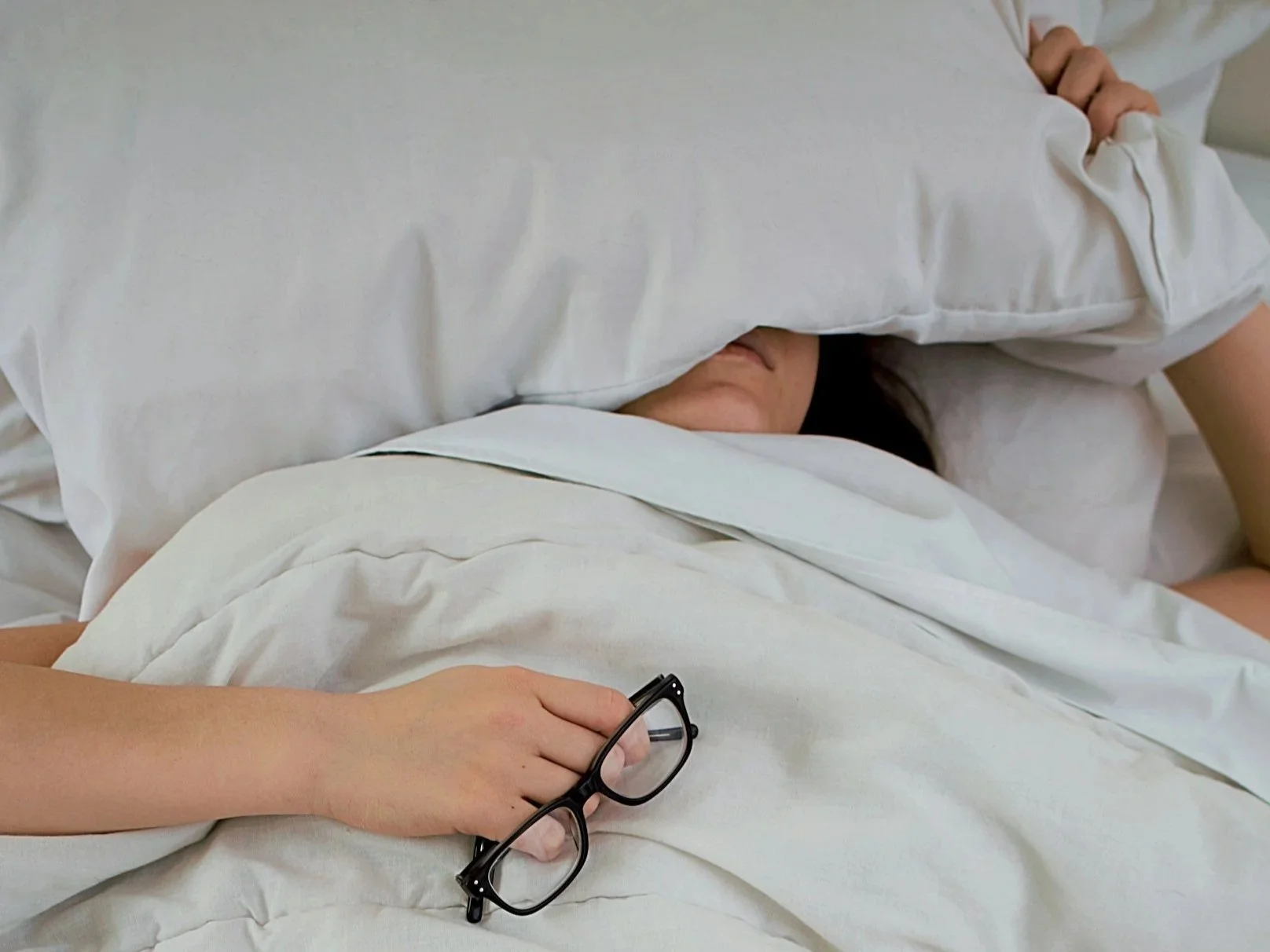 A woman attempting to sleep with insomnia in her white bedding and hold a pillow over head