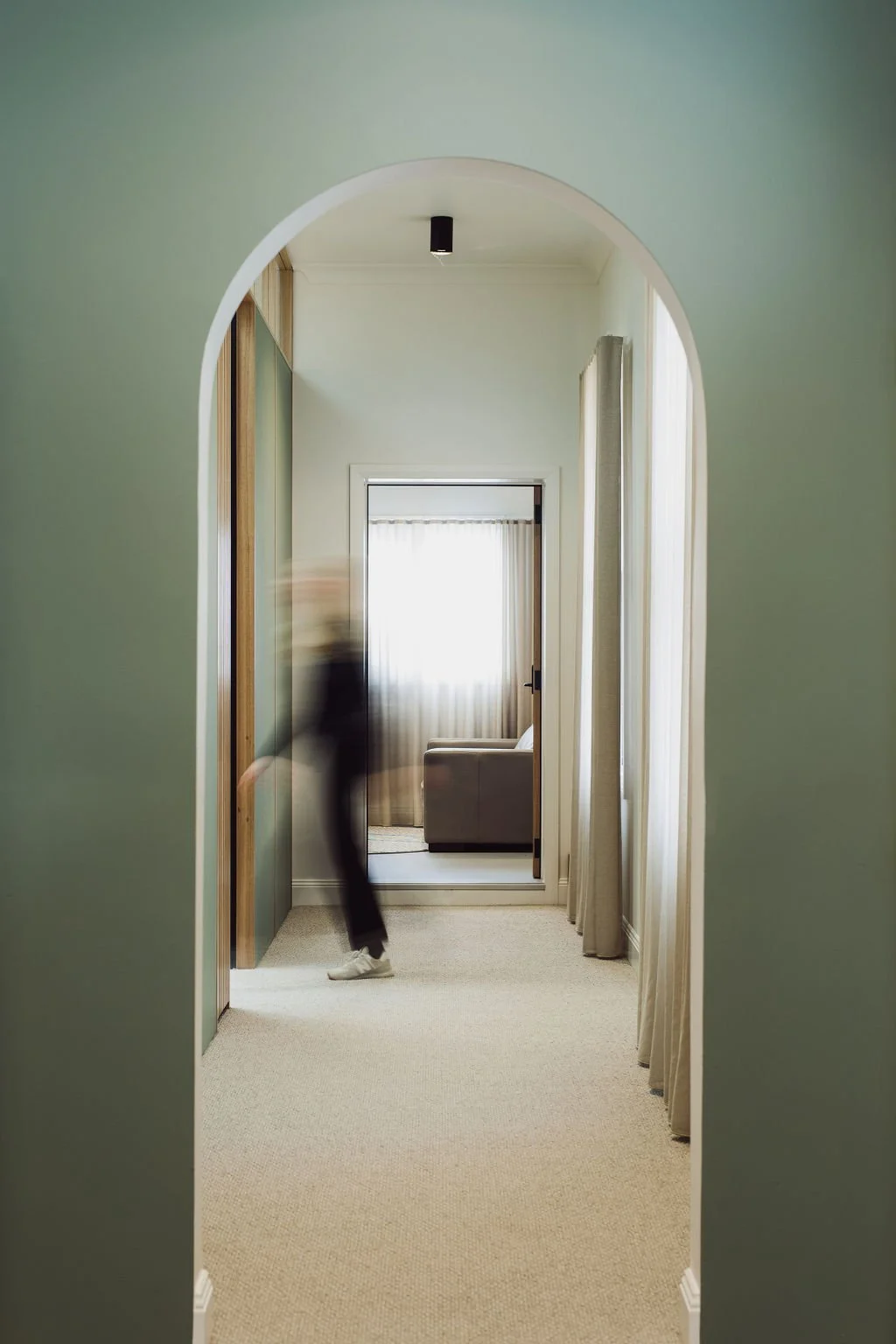 Blurred person walking through an arched doorway into a living room with beige carpet and a sofa.