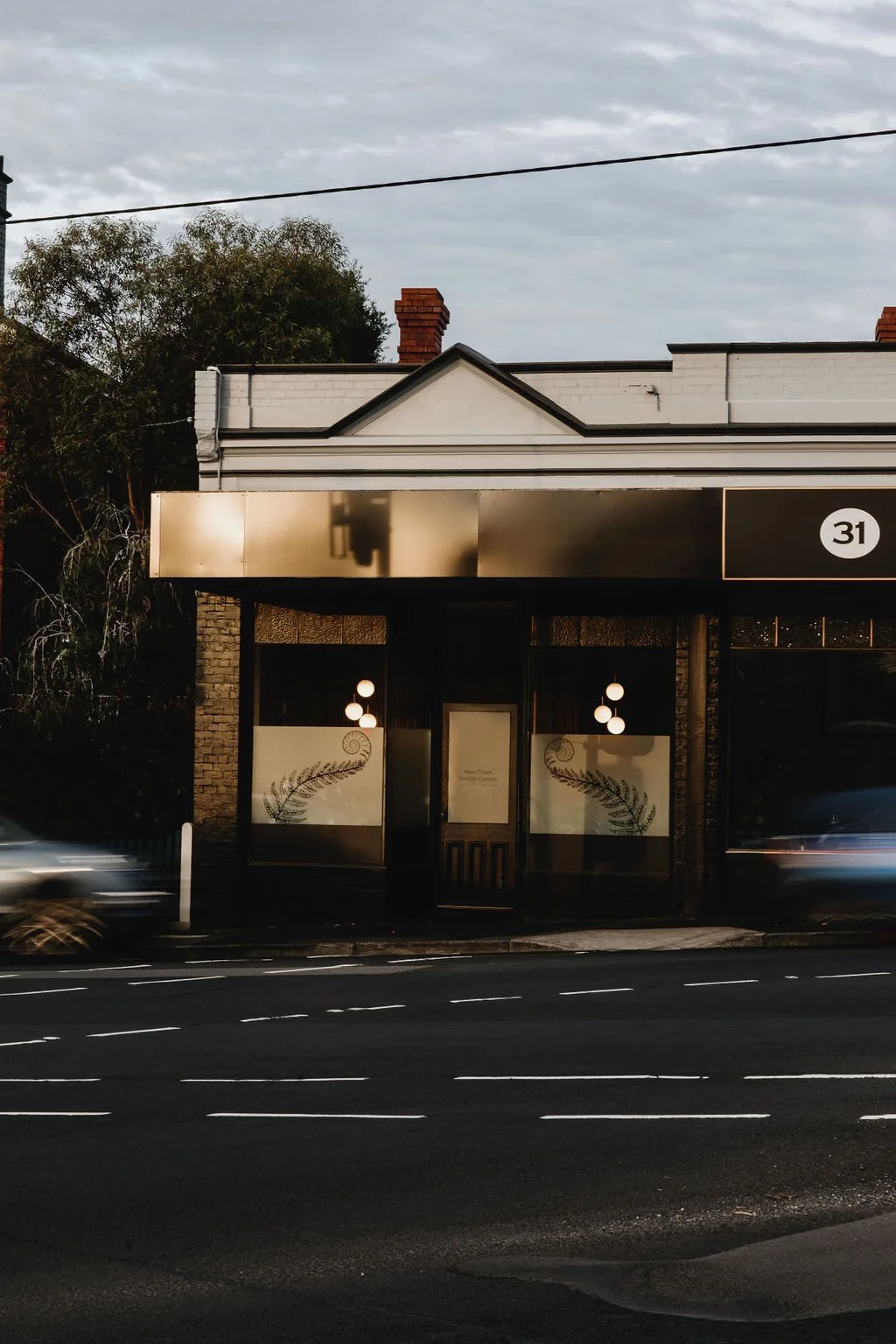 A storefront with illuminated windows and modern light fixtures outside, situated on a street with passing cars and a cloudy sky above.