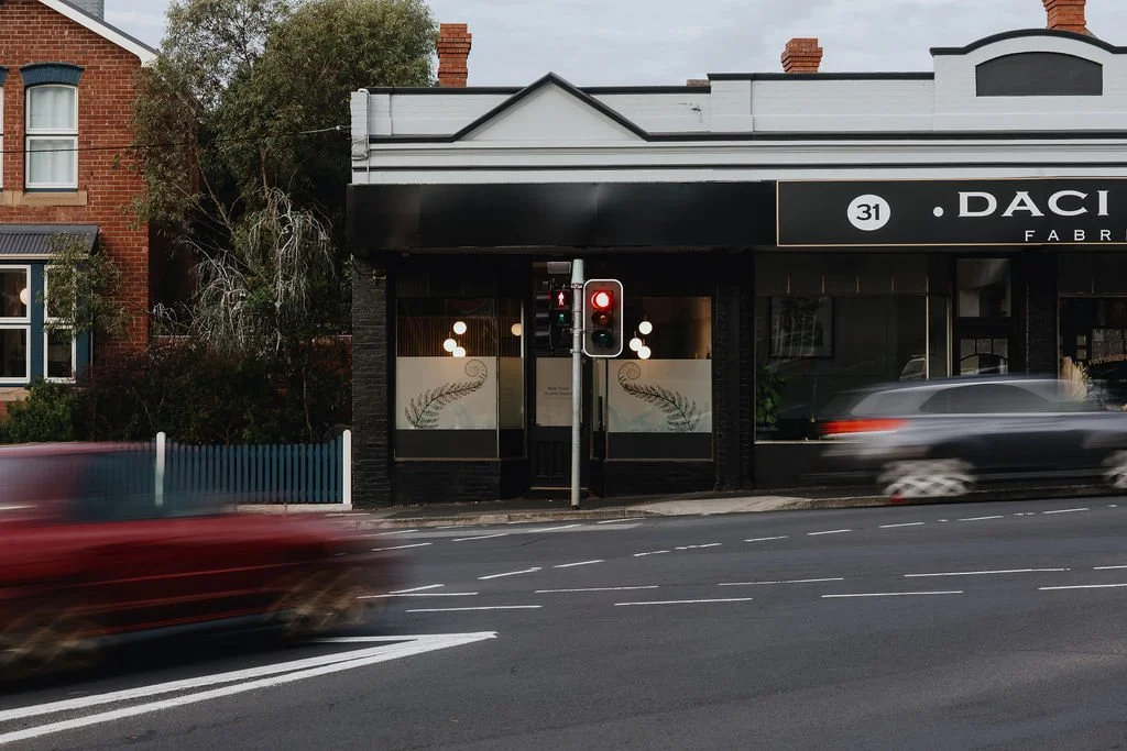 A street view showing a traffic light with a red signal, moving cars blurred in motion, a storefront with large windows, and a two-story building with a red brick facade to the left. The storefront has signage with the text '31 DACI' and a decorative