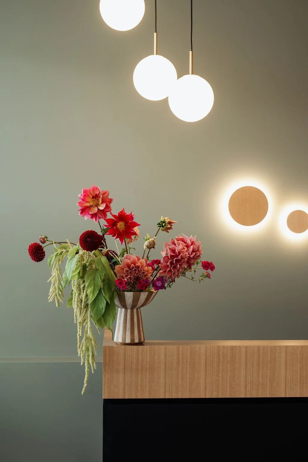 A bouquet of colorful flowers in a striped vase placed on a wooden surface, with modern hanging lights and circular wall lights in the background.