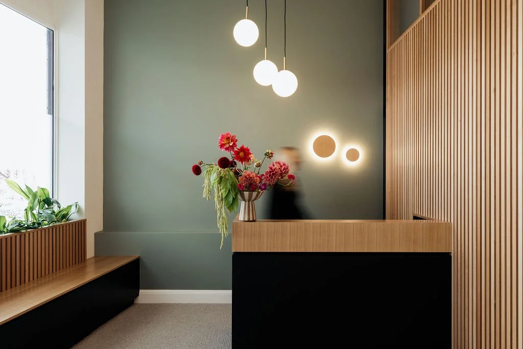 Modern reception area with a floral arrangement on the counter, wooden paneling, green accent wall, round wall lights, pendant lighting, and a window with plants.