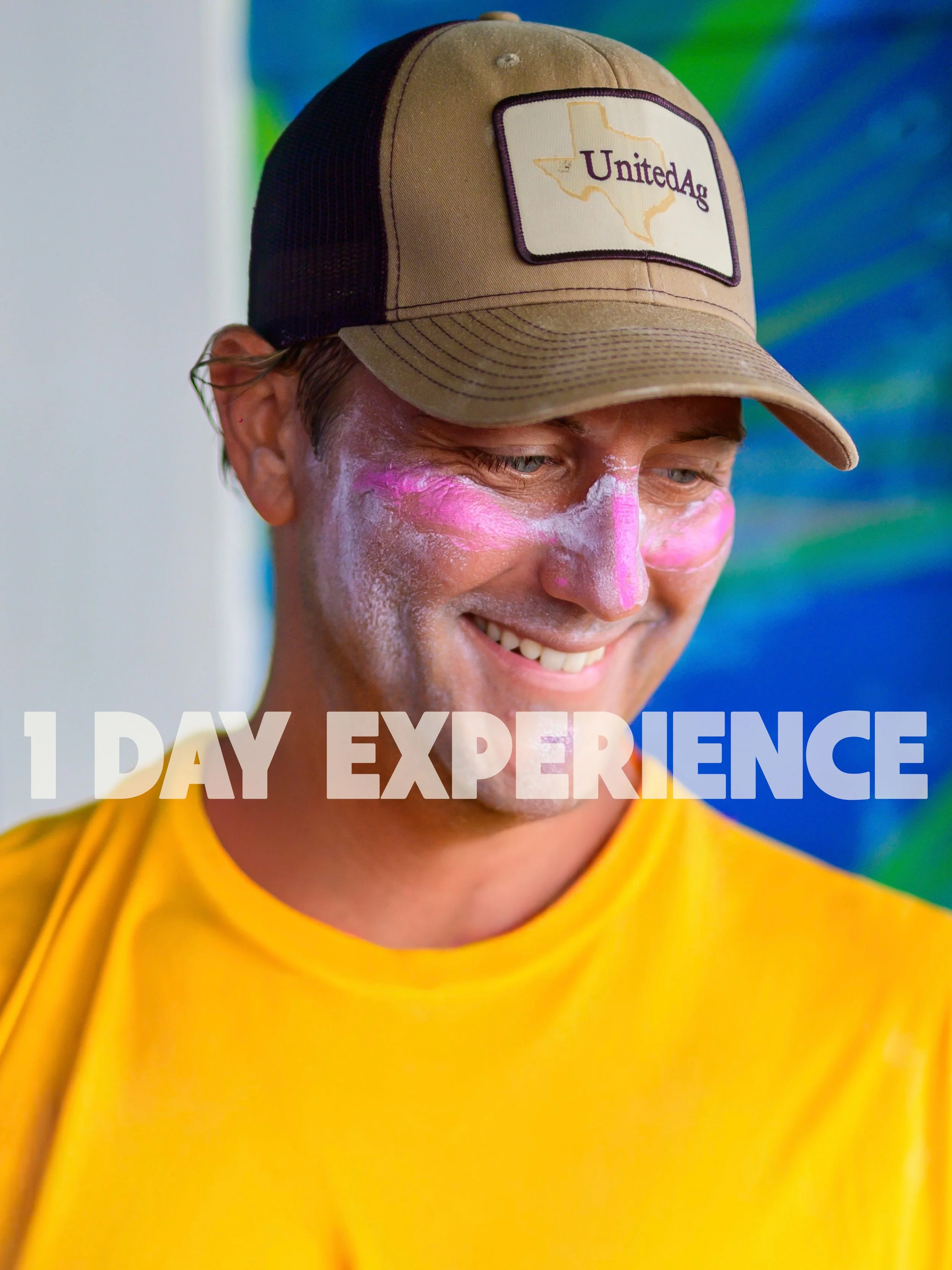 Smiling man with white and pink face paint wearing a tan and black cap with 'United Ag' logo and a yellow shirt, with a colorful background.