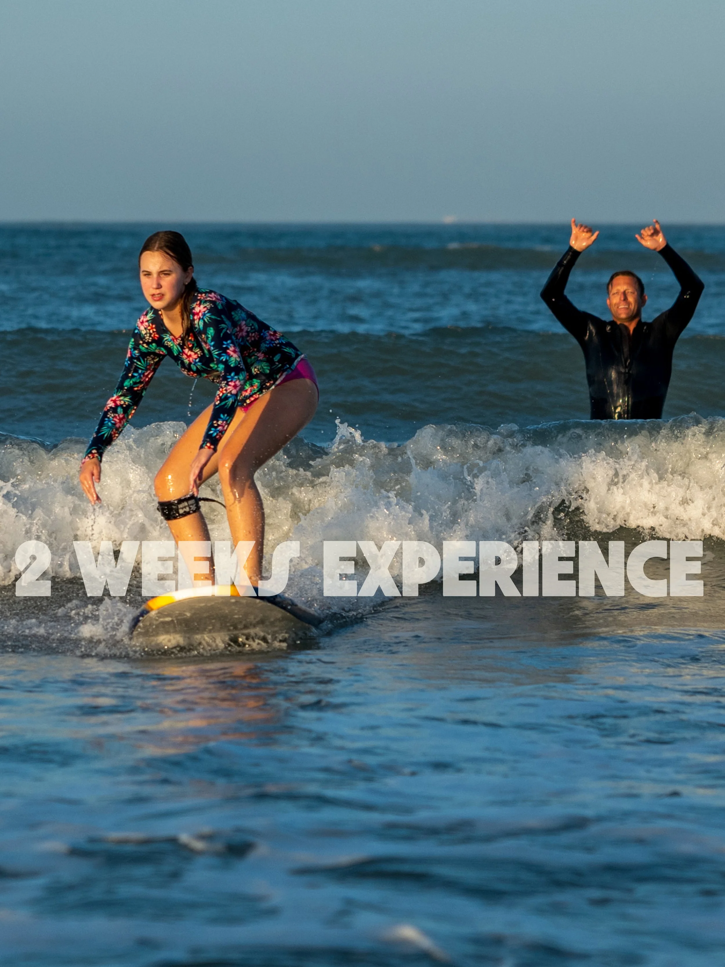 A young girl surfing on a wave with an adult man in the background celebrating in the ocean during sunset.