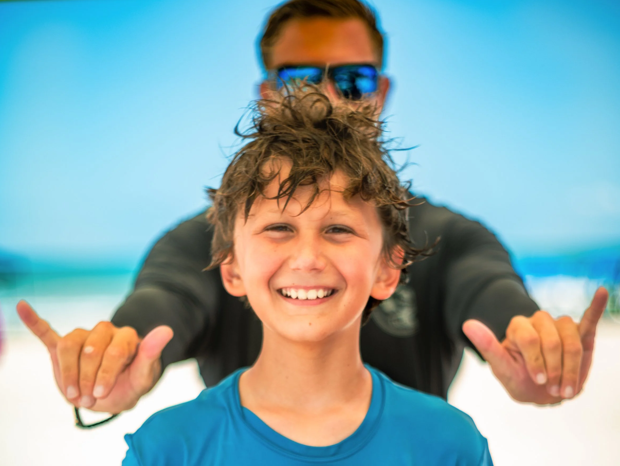 A smiling boy with wet hair in a blue shirt in front of a man with sunglasses making hand gestures at the beach.