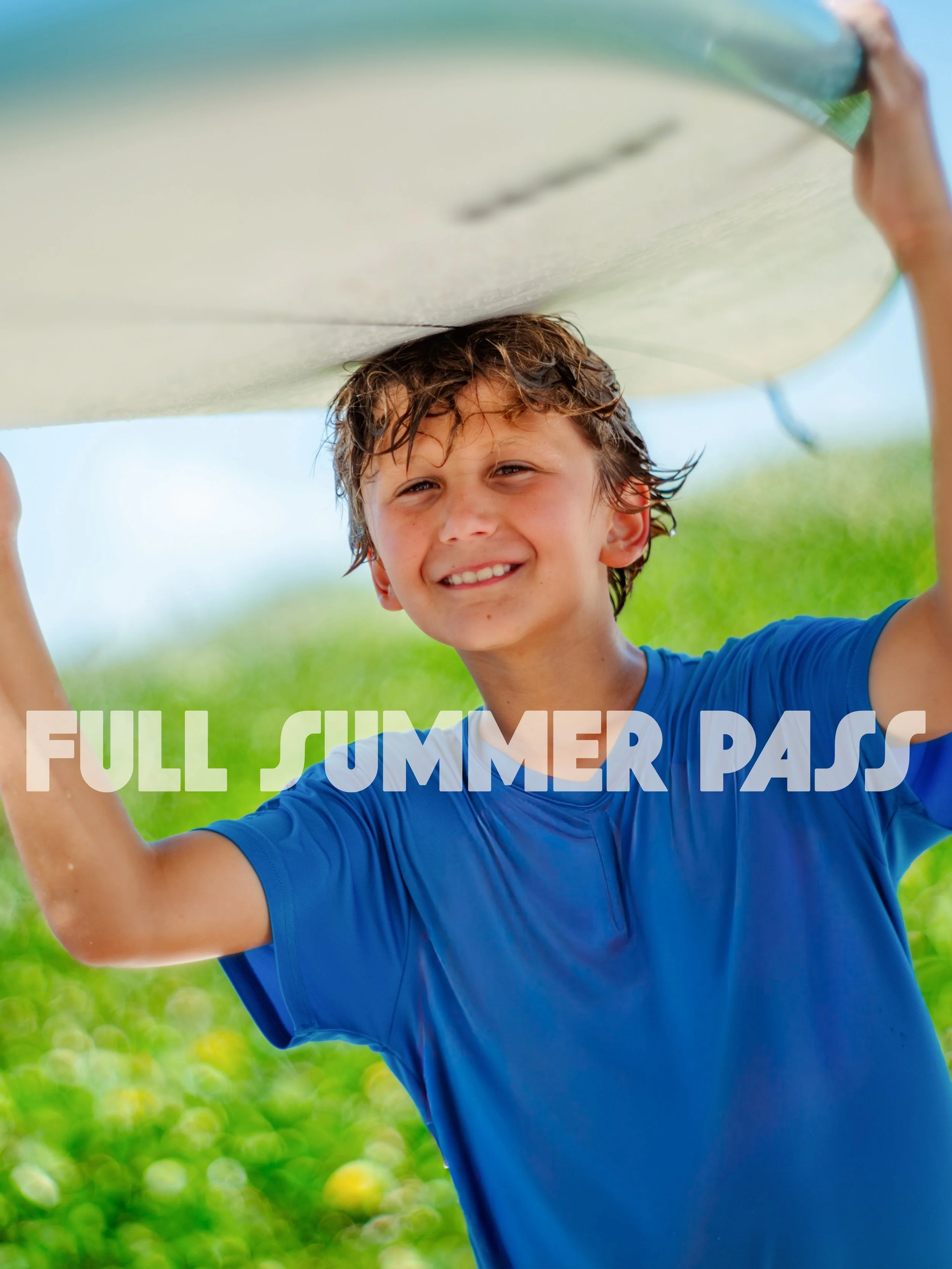 Young boy holding a surfboard overhead at the beach during summer, wearing a blue t-shirt, smiling, with green foliage in the background.
