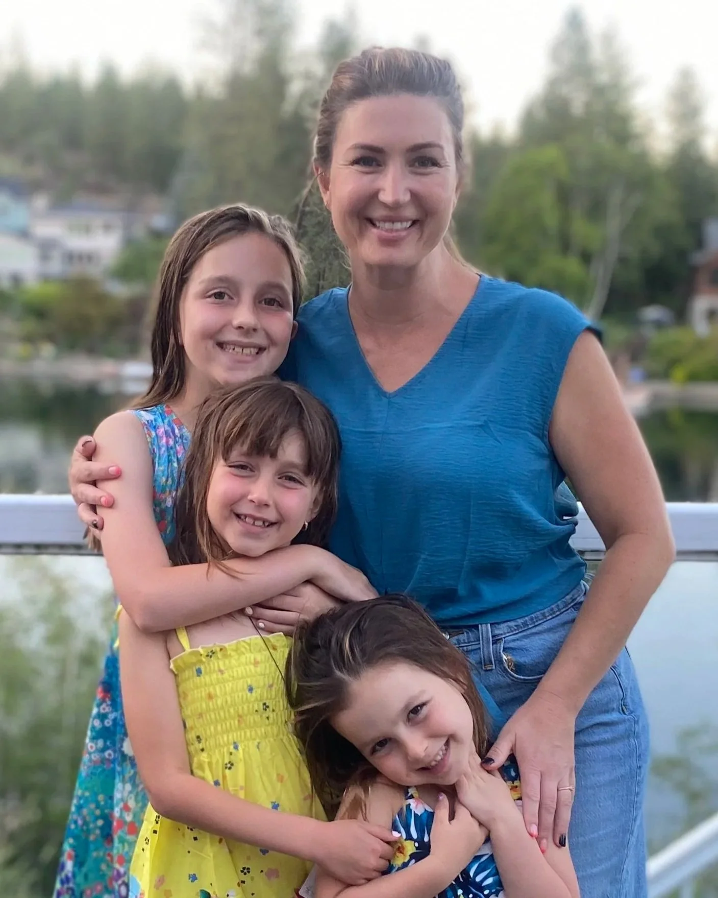 A woman and four young girls smiling and hugging outdoors near a lake, with trees and houses in the background.