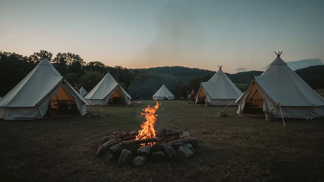 Several white canvas tents pitched across an open field during sunset or dusk, with a campfire in the foreground surrounded by rocks.