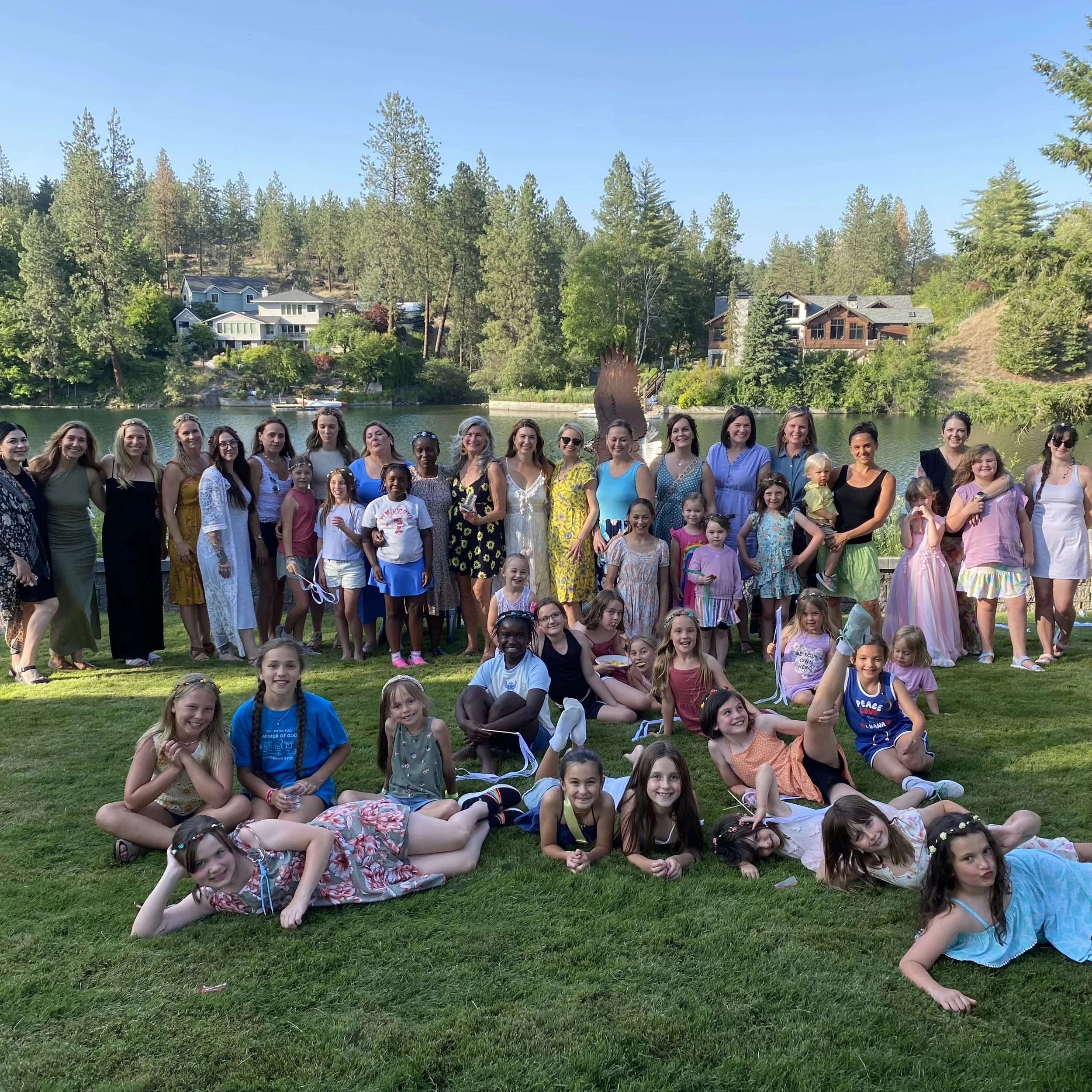 A large group of women and children posing for a photo outdoors near a lake with trees and houses in the background.