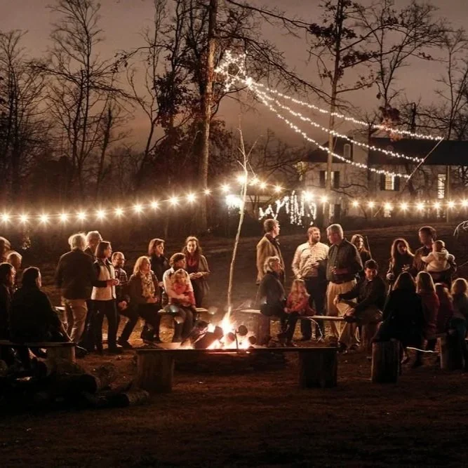 People gathered around a campfire outdoors at night with string lights overhead in a wooded area, celebrating an evening event.