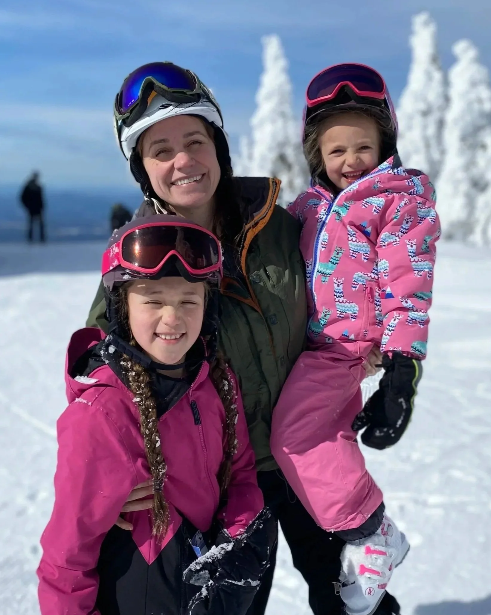 A woman and two young girls in pink ski jackets and snow gear posing together on a snowy mountain with snow-covered trees in the background.