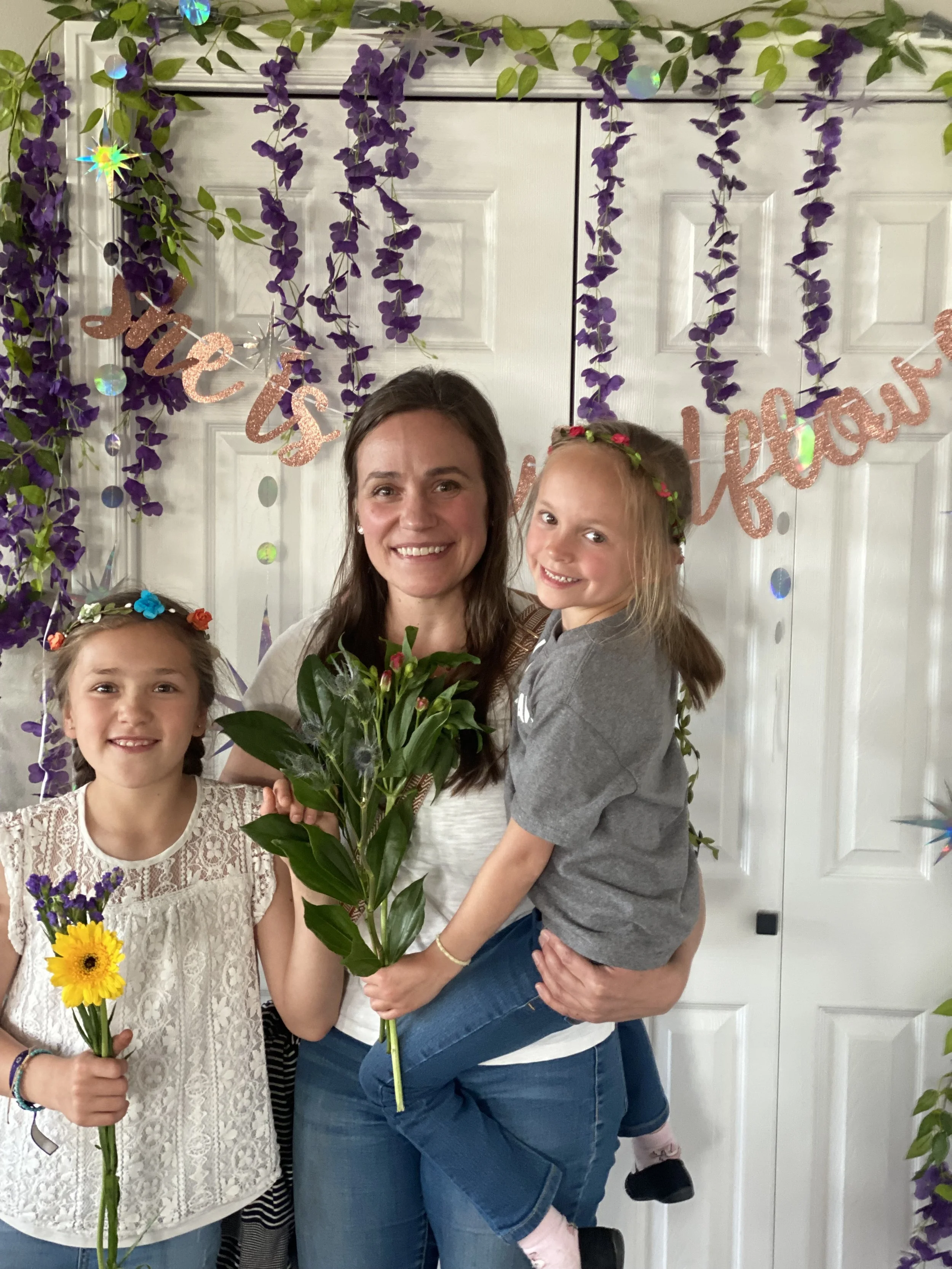 A woman with two young girls in front of a door decorated with purple flowers and a pink glittery banner that says 'it's a celebration'. The woman is holding a large green plant and the girl on the left has a yellow flower, while the girl on the righ
