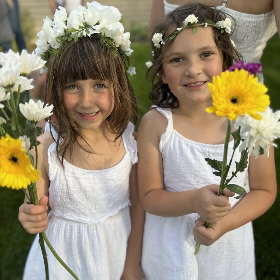 Two young girls wearing white dresses and floral crowns, holding yellow and white flowers, standing outdoors in a grassy area