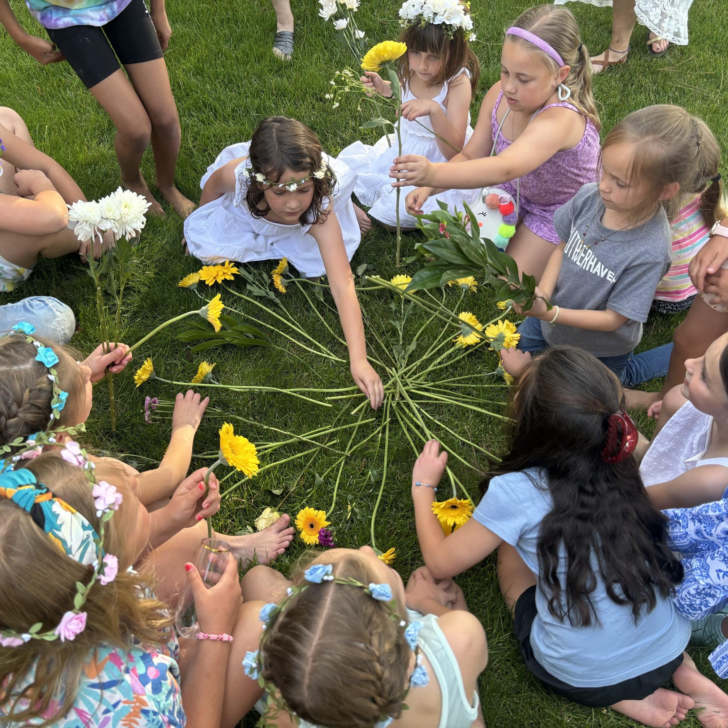 A group of children dressed in summer clothes and floral headbands are sitting and kneeling on grass, surrounding and arranging colorful flowers and greenery in a circular pattern on the ground. Some children appear to be adding flowers while others 
