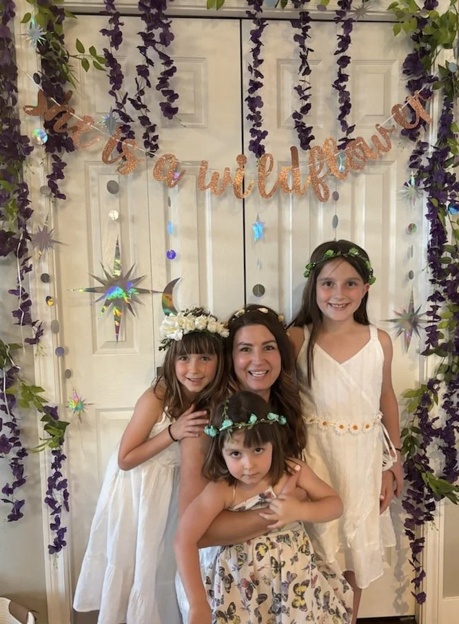 Four girls, including one adult woman, dressed in white and floral outfits, pose together in front of a decorated backdrop with purple flowers, stars, moons, and a banner reading 'It’s a Wildflower' in orange glittery letters.
