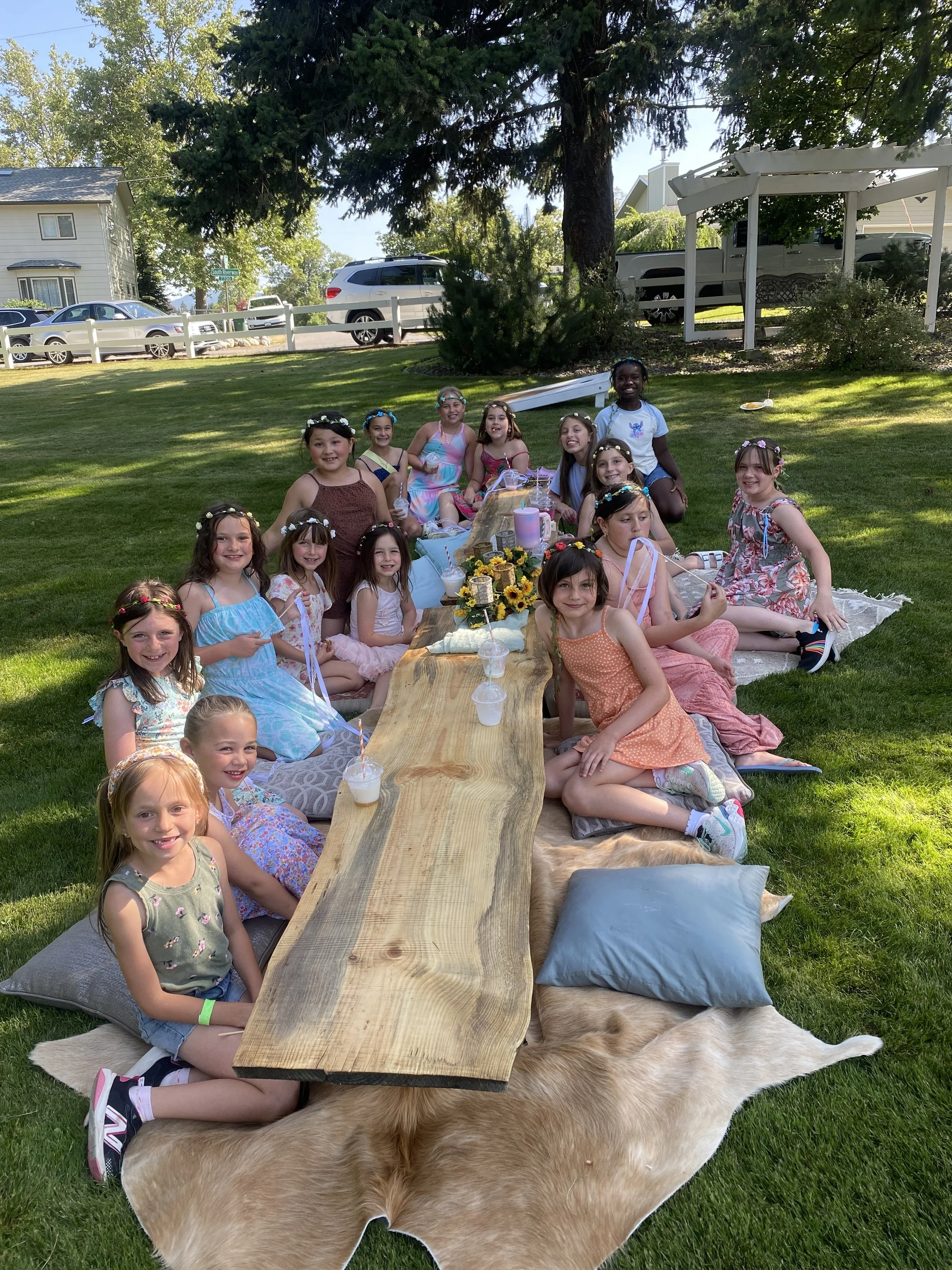 Kids at a birthday party sitting on cushions and a fur rug around a wooden table with drinks, celebrating outdoors on a sunny day.