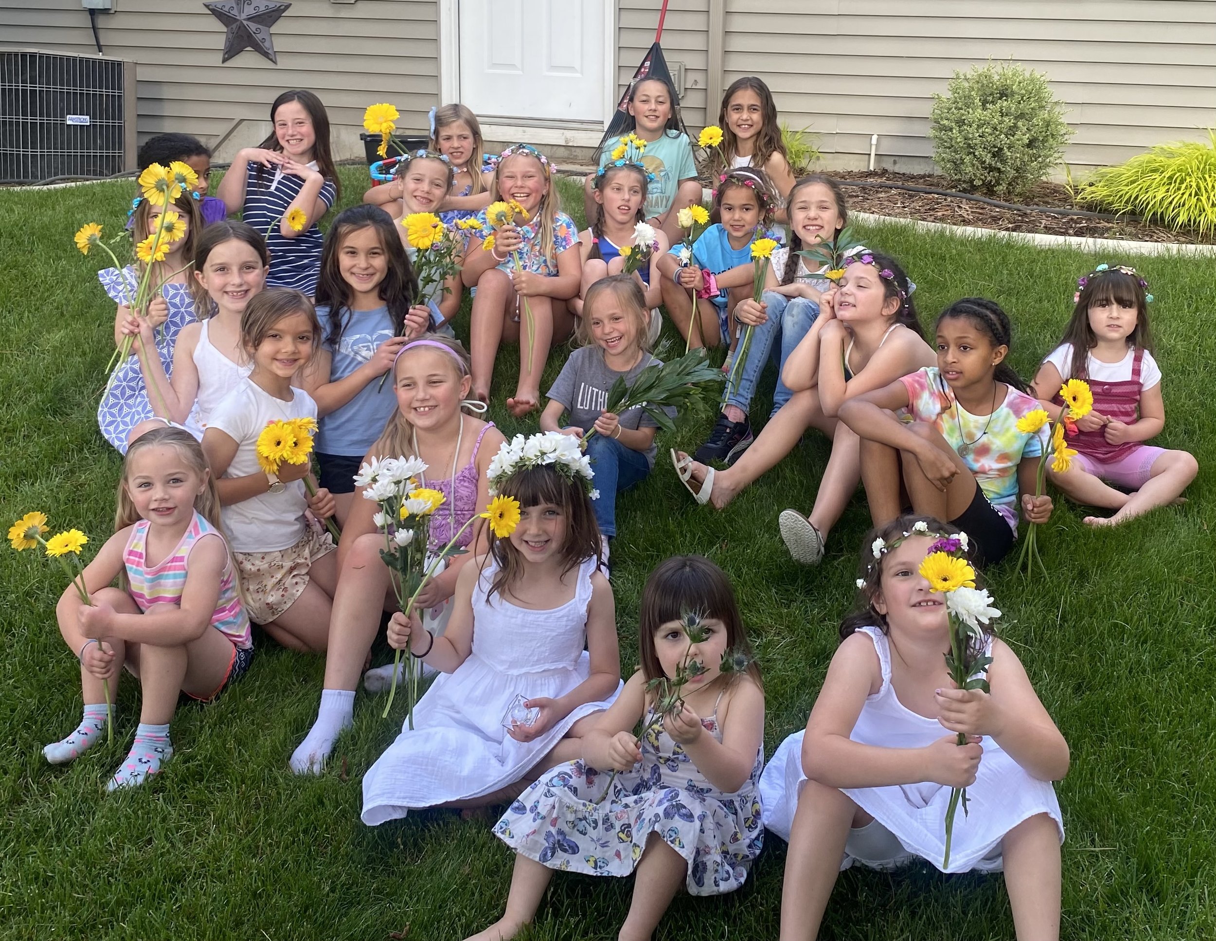 Group of young girls sitting and kneeling on a lawn in front of a house, holding yellow and white flowers, with some wearing flower crowns and colorful clothing, celebrating outdoors.
