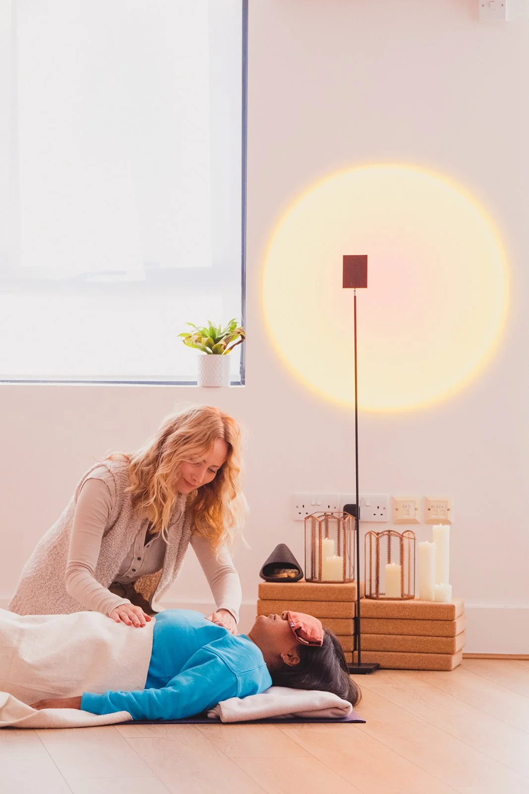 A woman lying down and another woman practicing Reiki or healing therapy in a peaceful room with soft lighting, candles, and decorative elements.