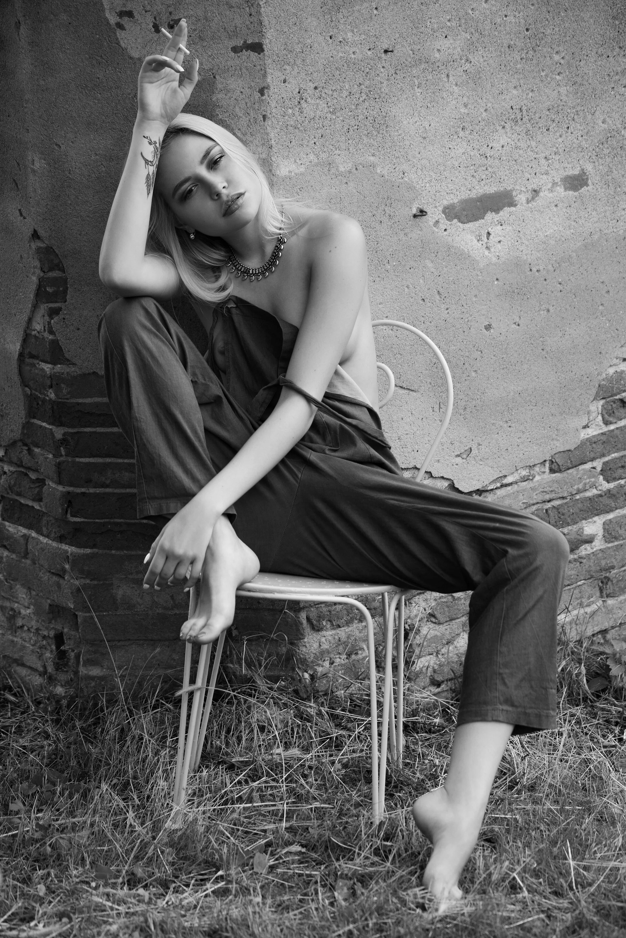 Femme nue assise sur une chaise en métal, posant contre un mur en briques et en béton, avec des herbes au sol, en noir et blanc.