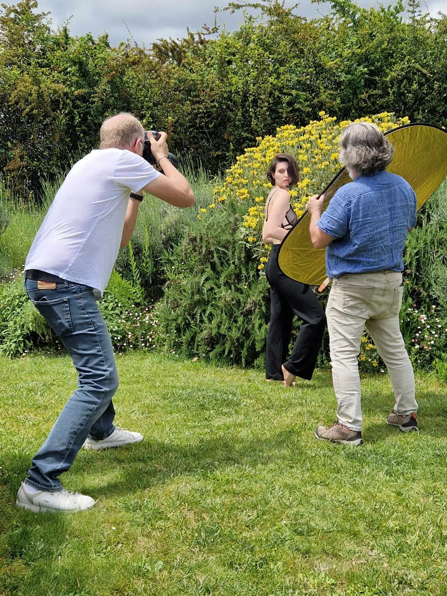 Une séance photo en plein air avec une femme modèle posant devant un buisson de fleurs jaunes, un photographe prenant des photos, et un assistant portant un réflecteur.