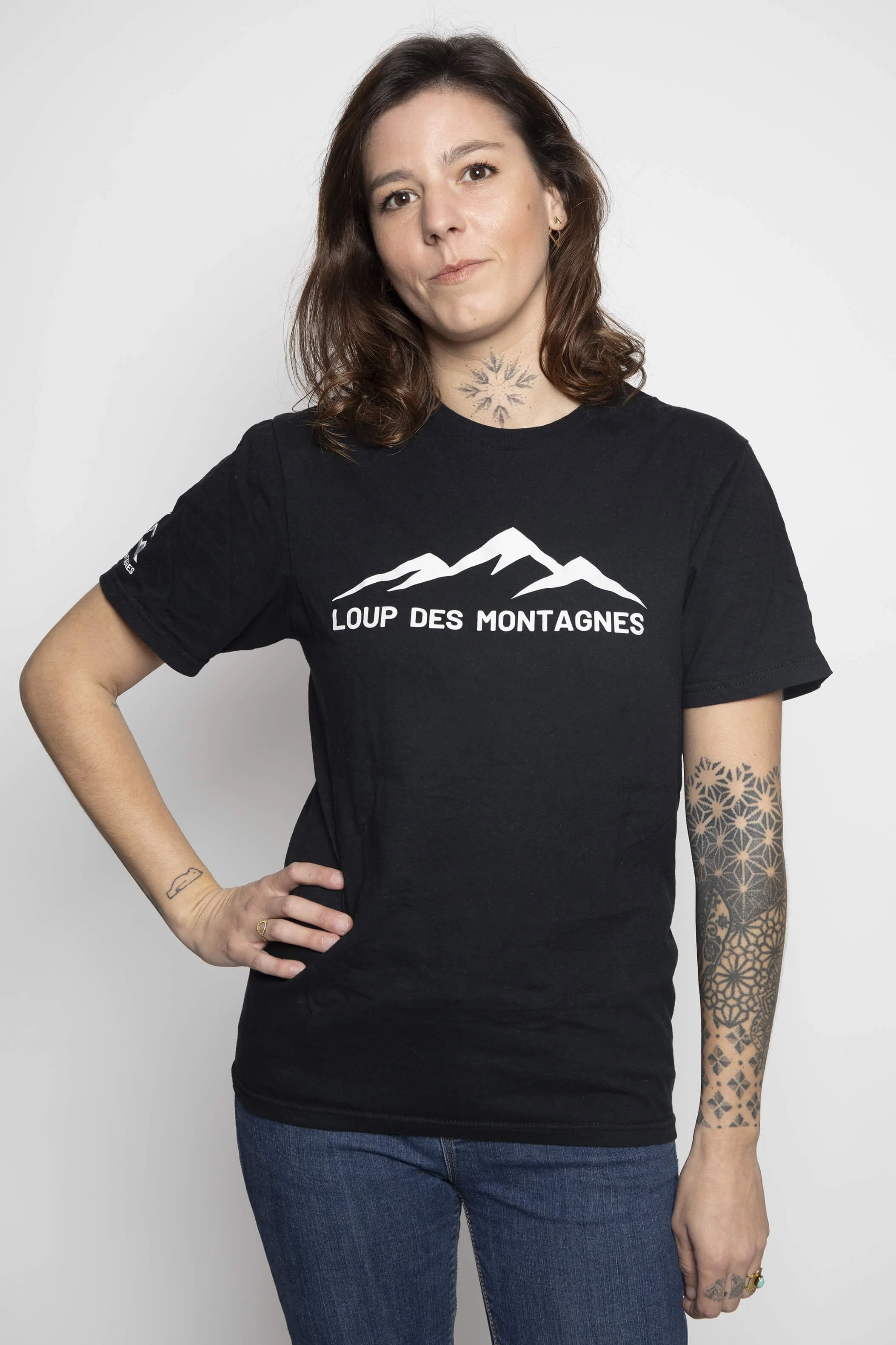 T-shirt noir avec le texte 'Loup des Montagnes'., porté par une femme avec des tatouages.