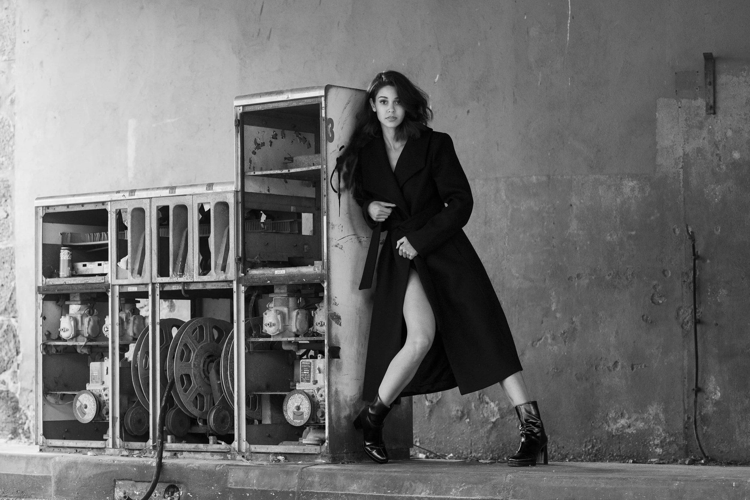 Une femme portant un manteau noir, debout contre une boîte électrique dans un environnement industriel, en noir et blanc.