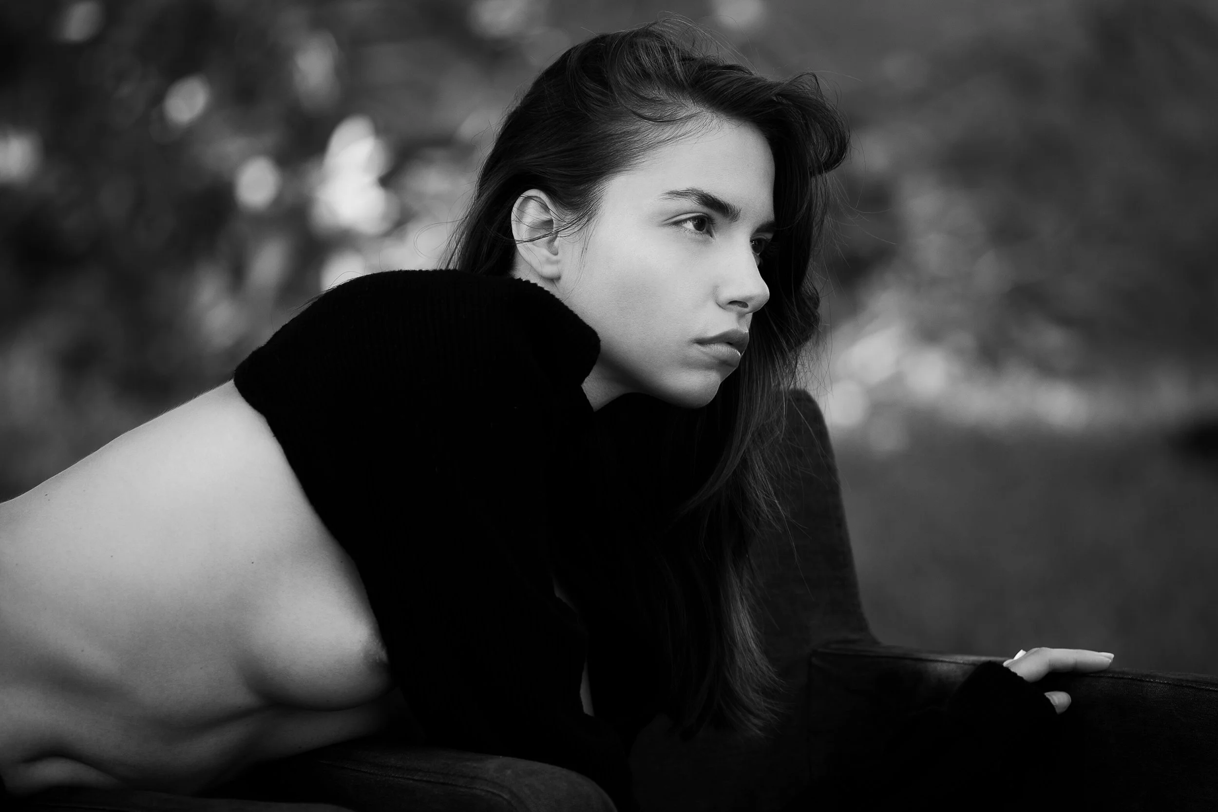 Une femme aux longs cheveux foncés, portant un haut sombre, est assise à l'extérieur, regardant pensivement vers la droite, avec un arrière-plan flou de nature.