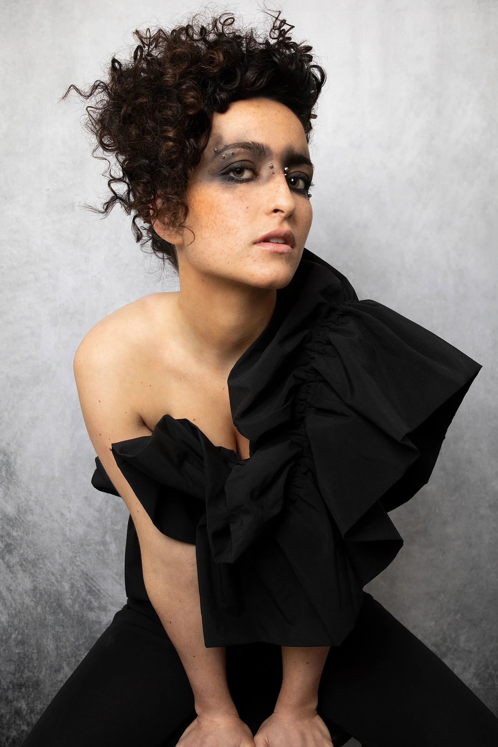 Portrait artistique d'une coiffure avec des boucles et du maquillage audacieux sur fond neutre.
Coiffure créative en noir avec une robe à volants et un décor minimaliste.
Portrait d'un modèle mettant en valeur une coiffure originale et un style de ma