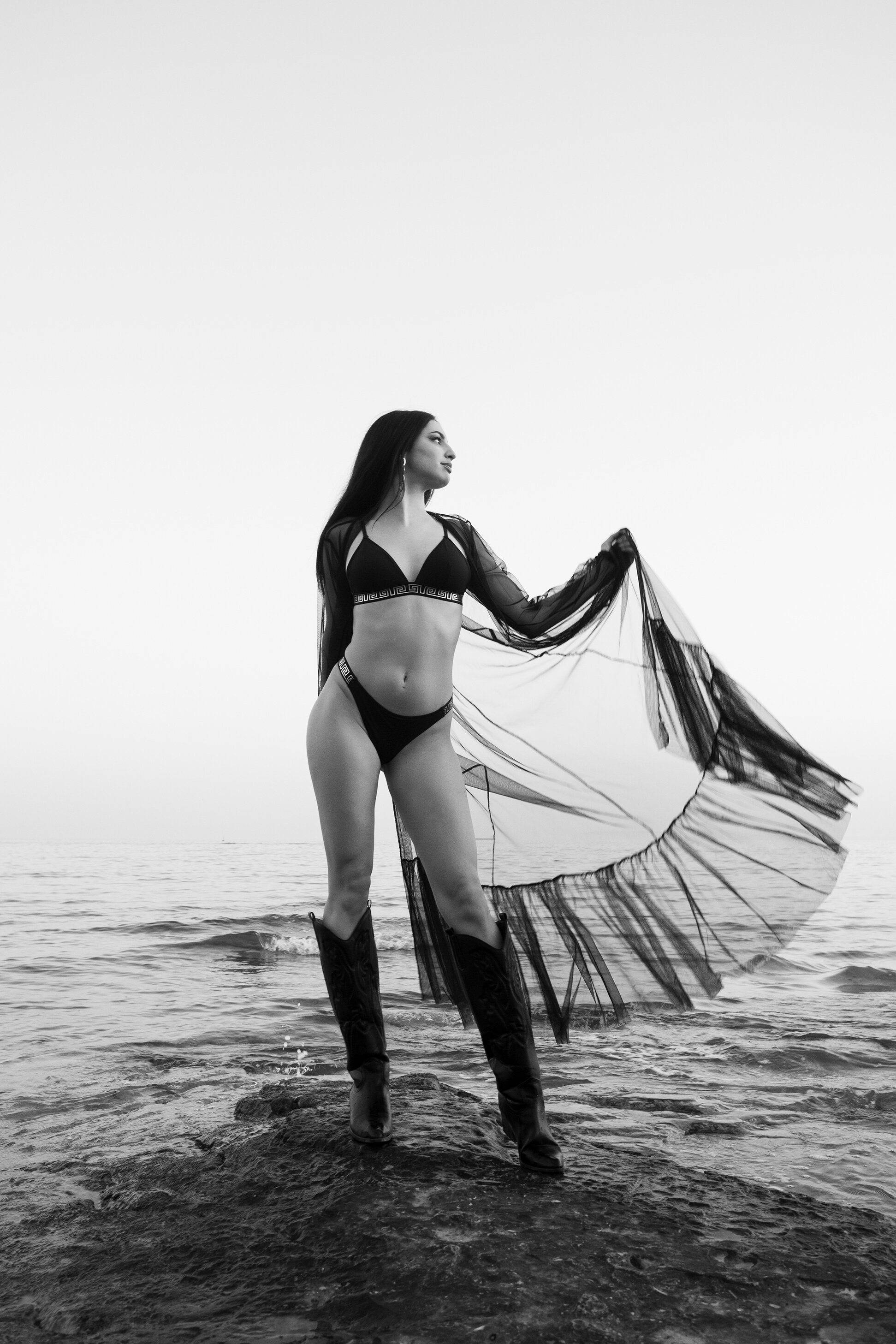 Une femme en maillot de bain noir, portant des bottes en caoutchouc, se tient sur une roche à la plage, tenant un voile noir transparent qui se déploie dans le vent, photographiée en noir et blanc.