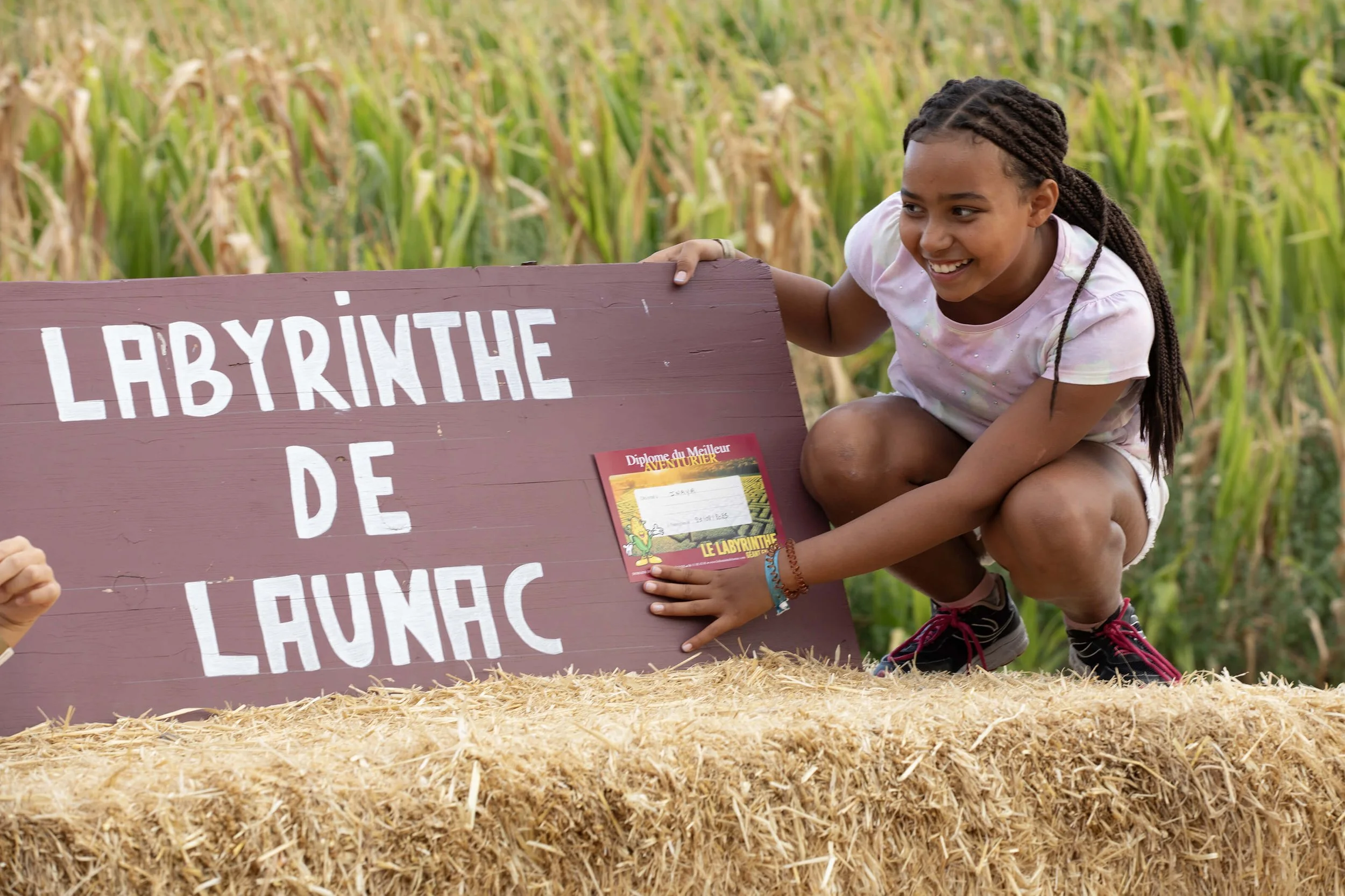 Un enfant souriant pose fièrement près d’un panneau au labyrinthe de Launac, un diplôme à la main.  La photographie capture l’ambiance joyeuse du décor champêtre du labyrinthe, avec la jeune fille rayonnante de bonheur.  Les couleurs vives du panneau
