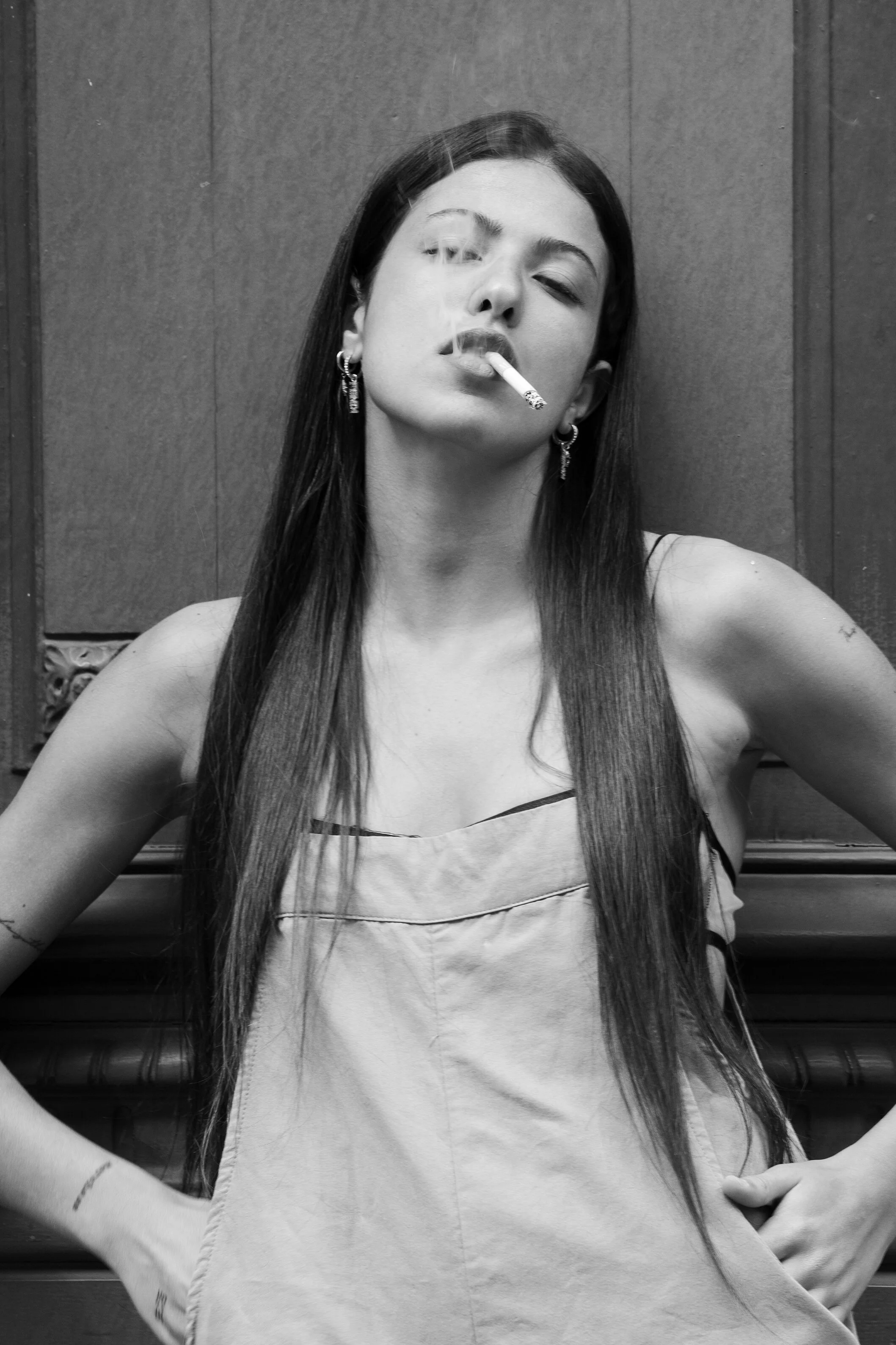 Une jeune femme avec de longs cheveux noirs porte une robe sans manches et fume une cigarette, devant un mur en bois.