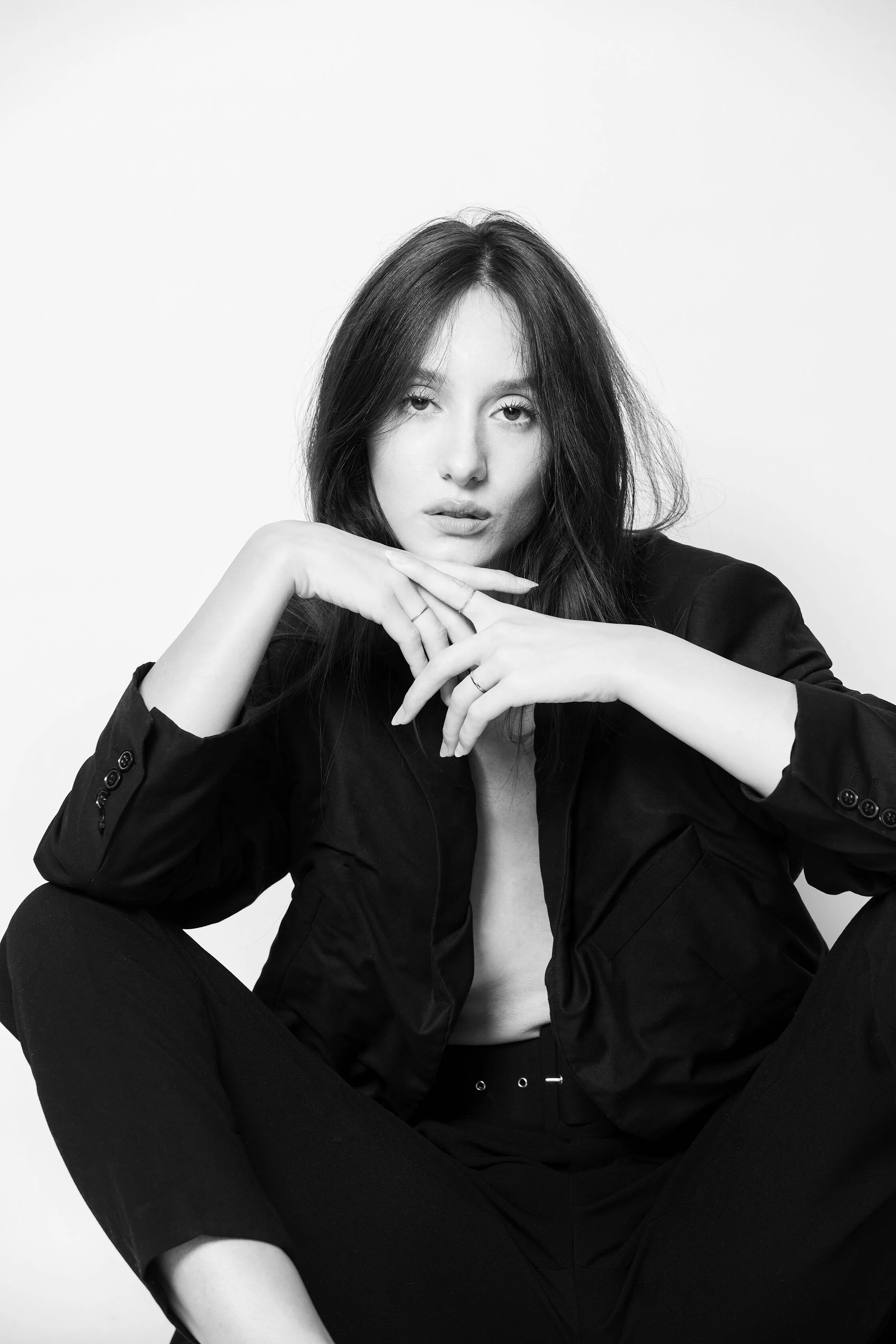 Portrait en noir et blanc d'une femme avec cheveux longs, portant une veste noire, assise en position décontractée, regardant droit devant, fond blanc.