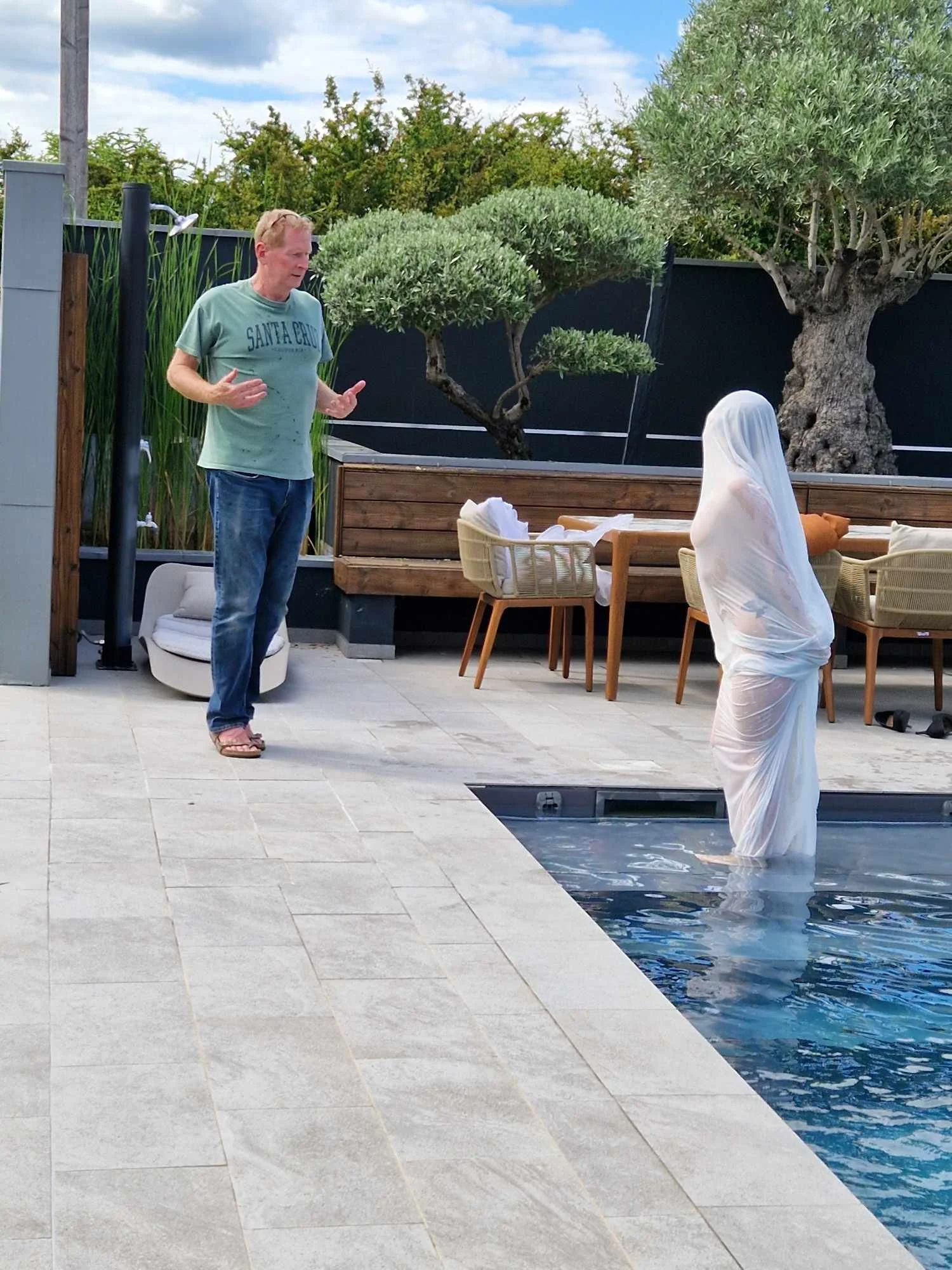 Un mannequin portant un drapé blanc ressemble à une statue dans une piscine, face à François Fauré qui parle, dans un environnement extérieur avec des arbres et un mobilier de jardin.