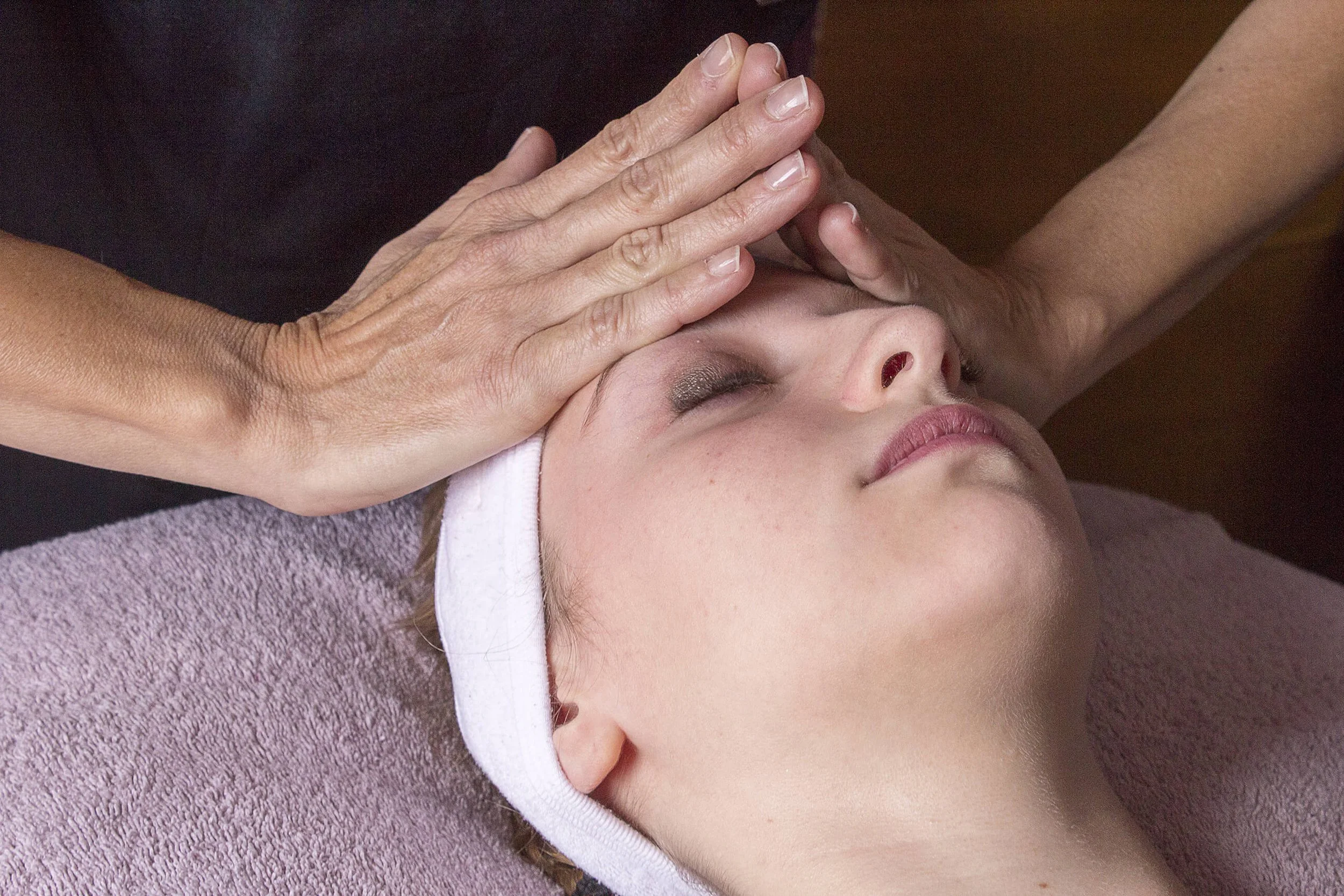 Dans un cadre professionnel, une femme reçoit un soin du visage, mettant en avant les mains expertes de l’esthéticien.  Un massage facial relaxant est illustré par un détail des mains, avec des couleurs douces et apaisantes. L’ambiance élégante d’un 