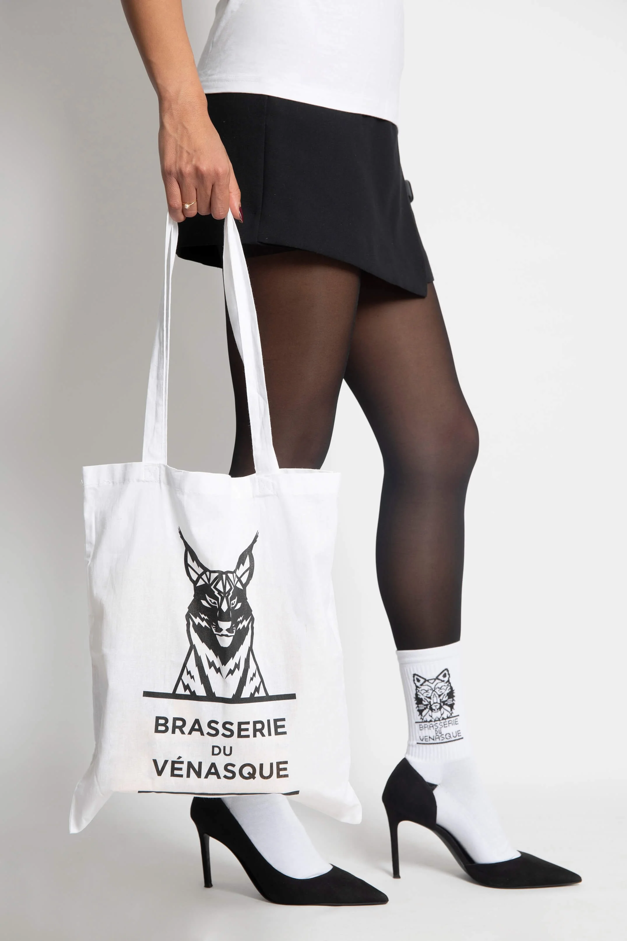 Femme en jupe noire et haut blanc portant un sac en toile et des chaussettes blanches avec le logo de la brasserie du Vénasque