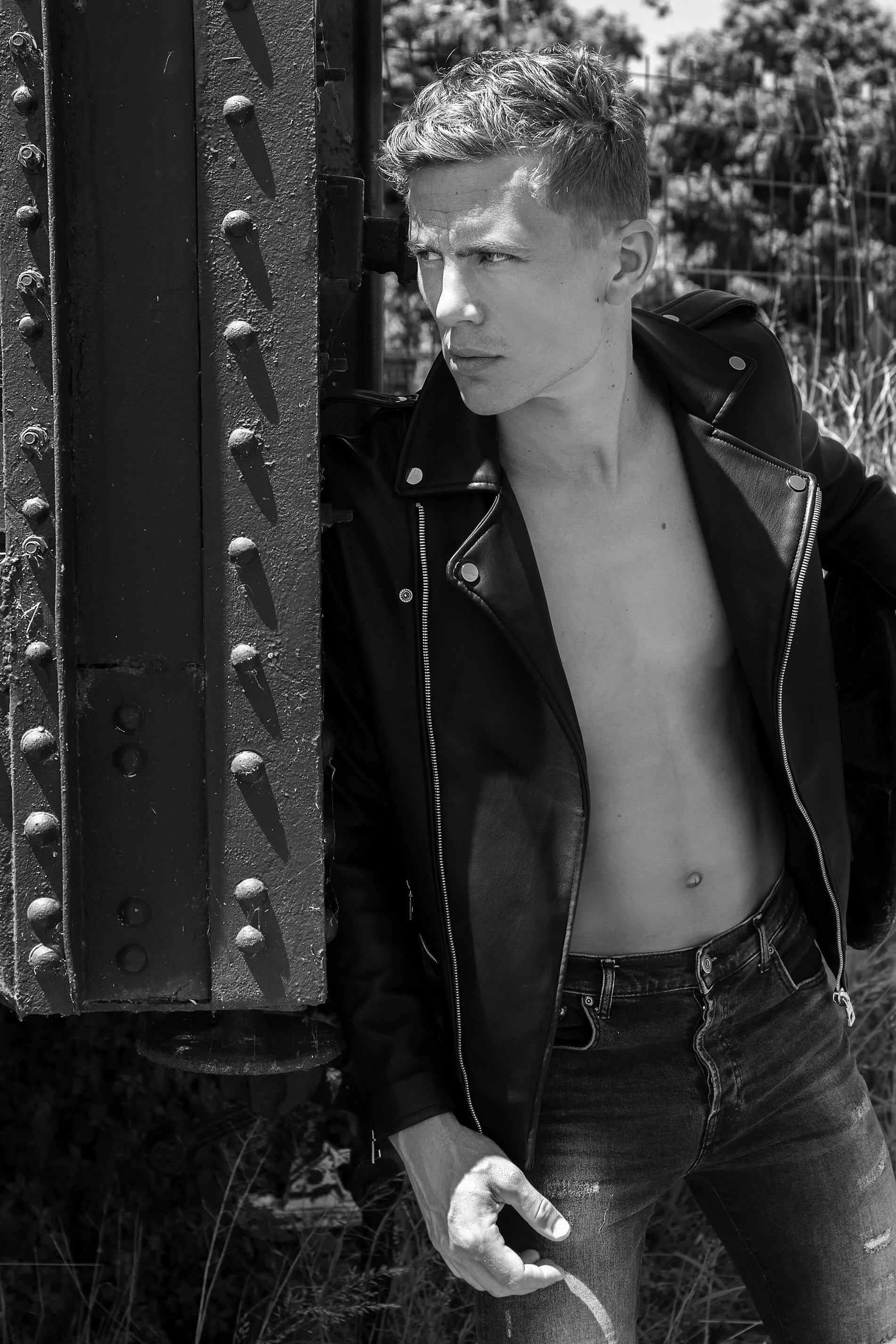 Un homme en jean noir, avec une veste en cuir ouverte, sans chemise, pose près d'une structure métallique, regardant vers la droite.