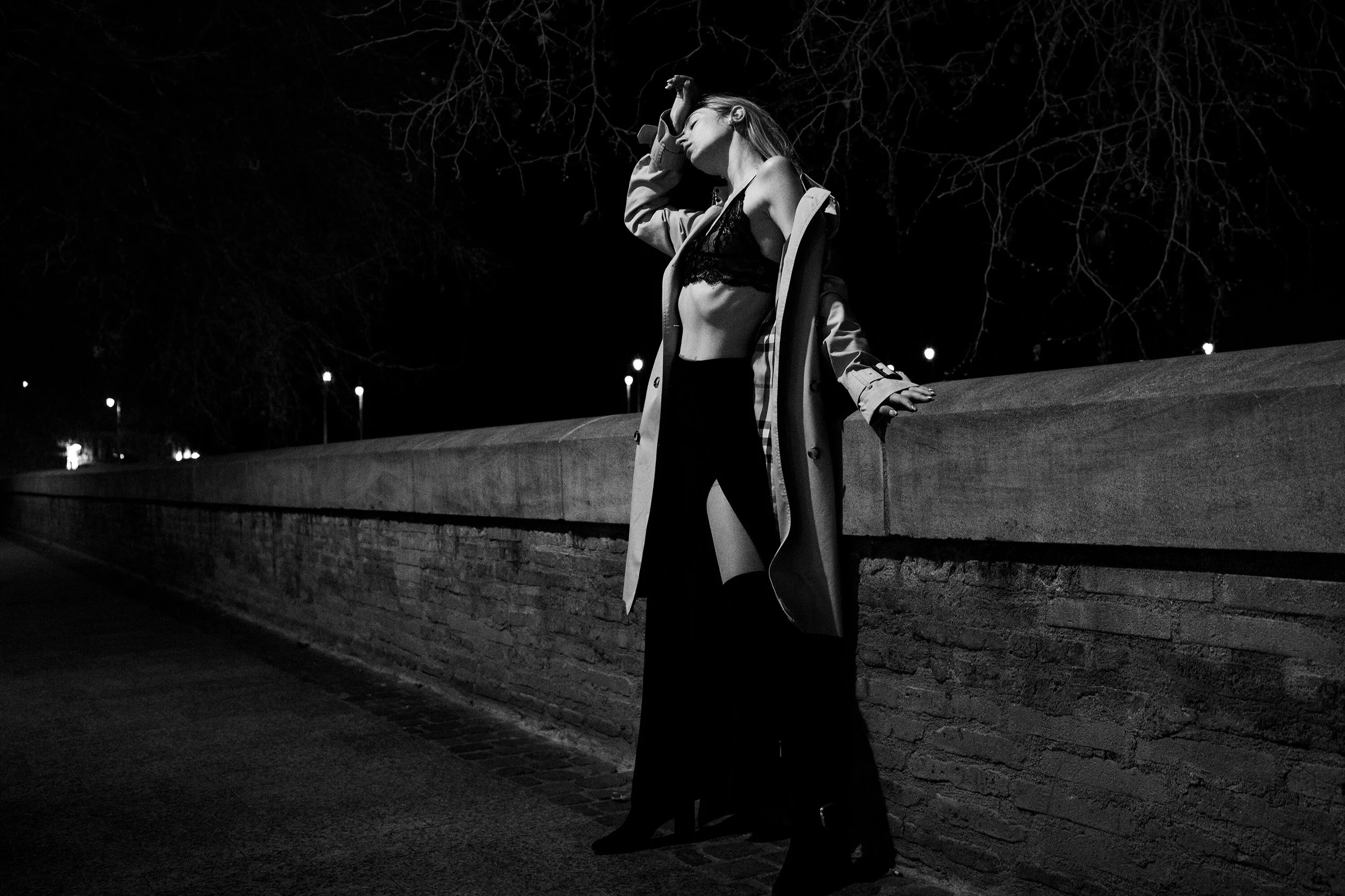 Femme portant un trench et une robe en dentelle, debout contre un mur en pierre la nuit.