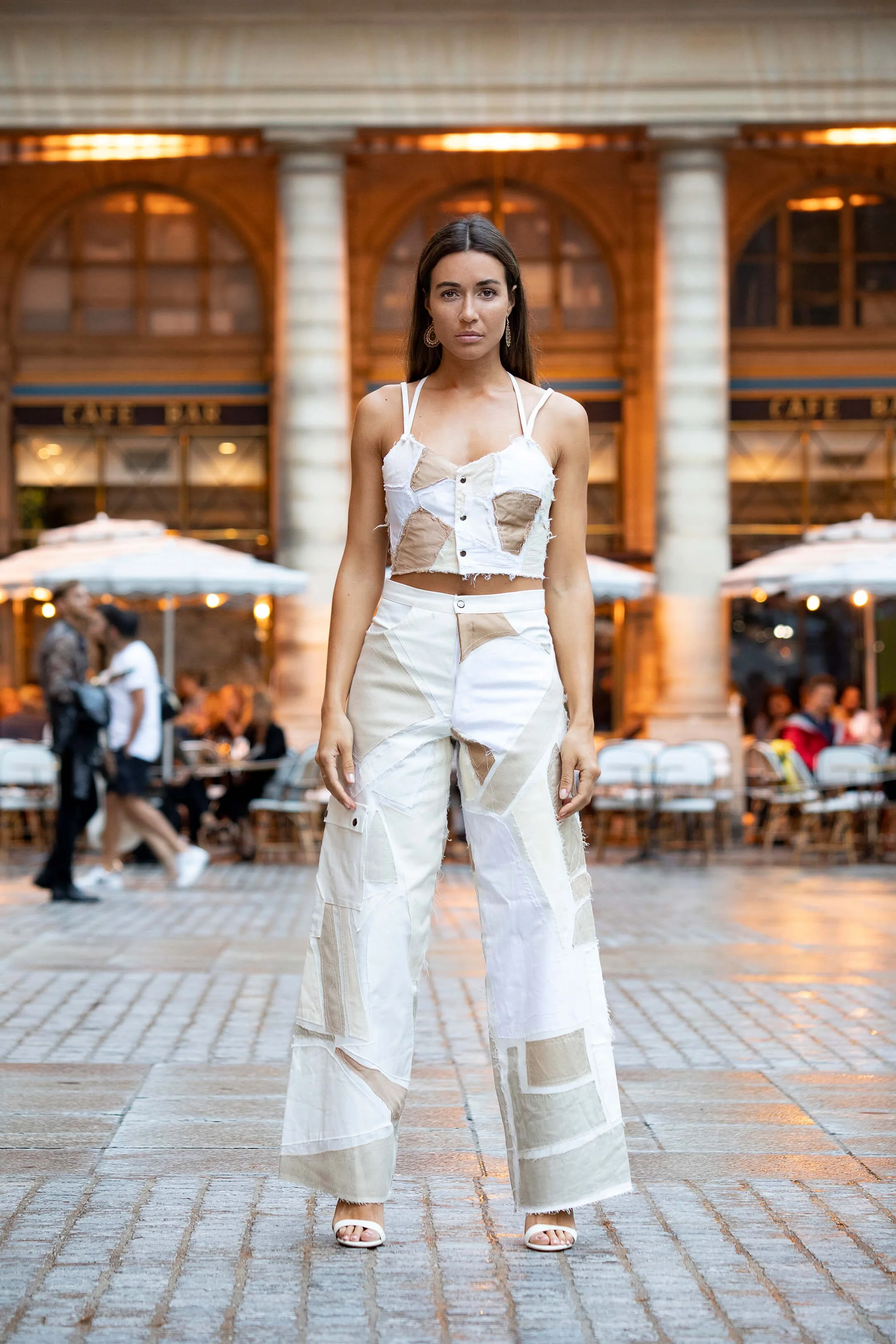 Femme portant un pantalon blanc unique avec des détails en patchwork dans un cadre café animé avec des clients en arrière-plan