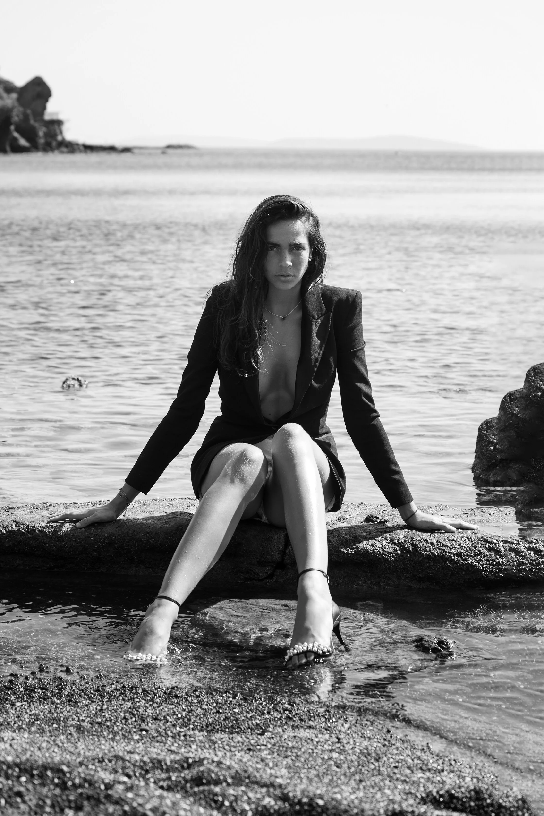 Une femme habillée en blazer noir, assise sur un rocher au bord de l'eau, en noir et blanc.