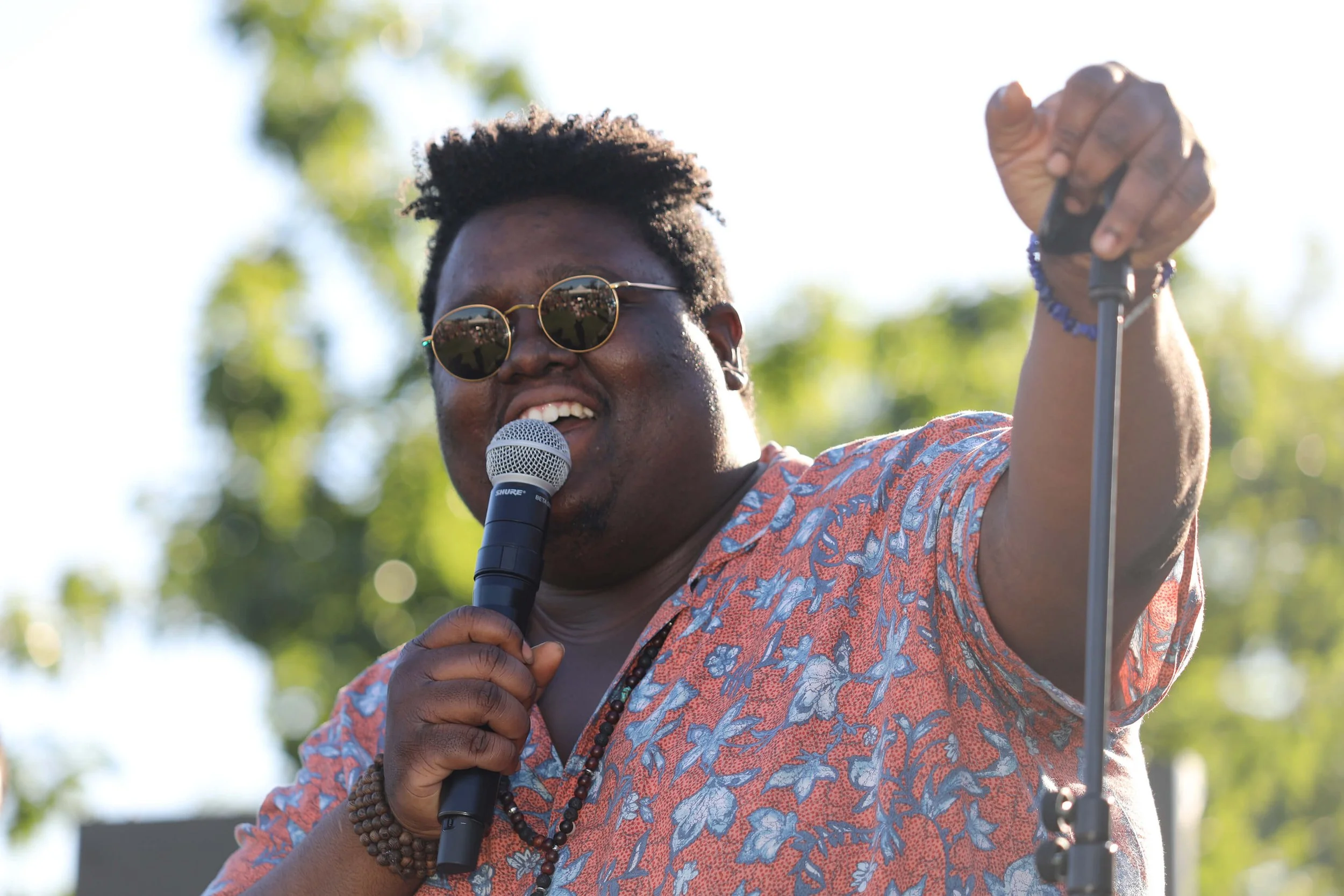 Un artiste souriant sur scène, micro à la main, capture l’énergie vibrante d’un événement en plein air.  Vêtu d’une chemise colorée, il interagit avec le public, créant une ambiance festive.  Le photographe François Fauré immortalise ces moments uniq