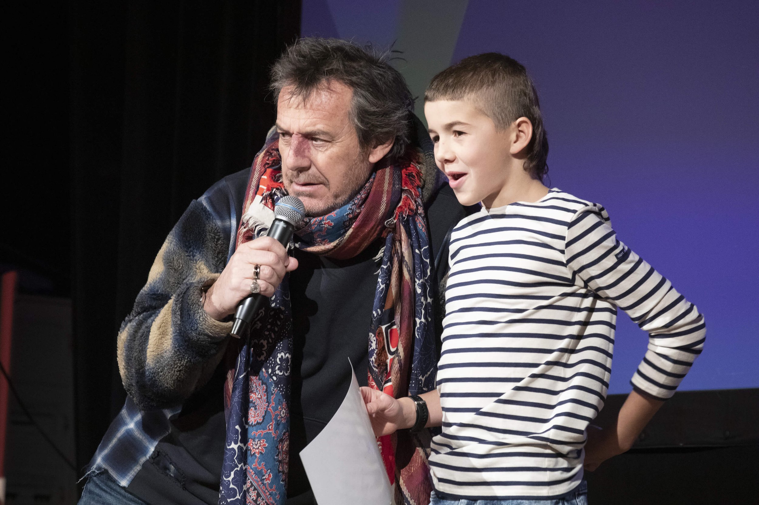 Jean-Luc Reichmann partage un moment chaleureux et enthousiaste avec un jeune participant lors d’un événement.  Le style photographique met en valeur l’interaction entre l’animateur et l’enfant sur scène.  François Fauré capture l’ambiance colorée et