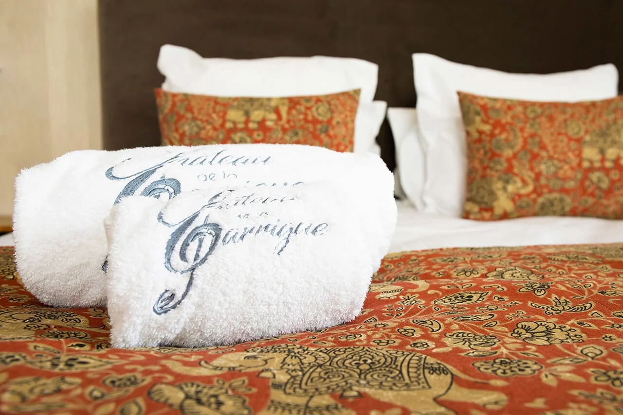 Des serviettes blanches ornées d’un logo reposent sur un lit décoré de coussins colorés.  L’intérieur de la chambre est sublimé par des draps et des coussins élégants.  Le photographe François Fauré nous offre une vue artistique d’une chambre d’hôtel